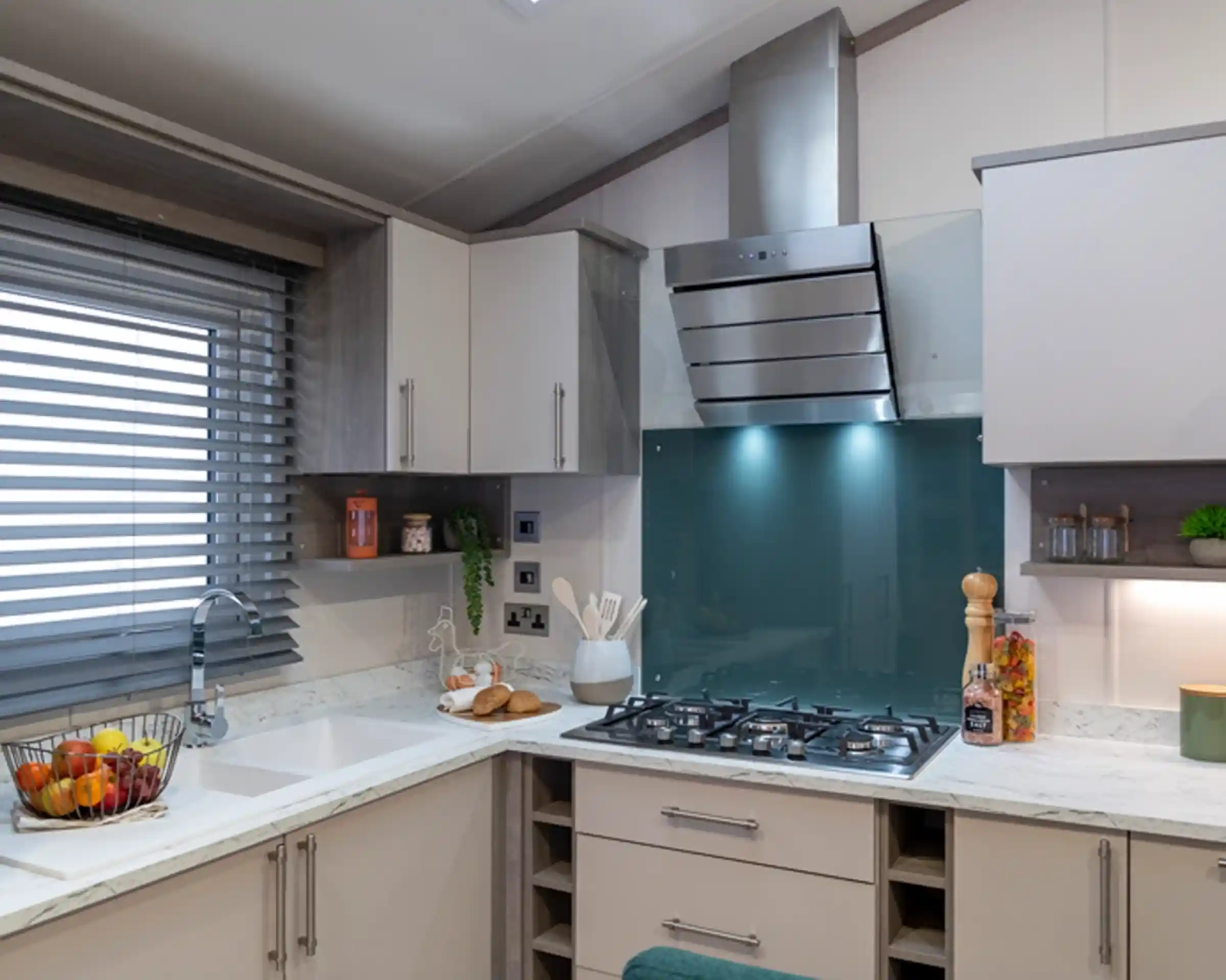 Contemporary kitchen with light cabinetry, a white marble-style countertop, and a teal glass backsplash behind a stainless steel gas cooktop and extractor hood. The kitchen includes a built-in sink below a window with modern horizontal blinds, open shelving with decorative items, and various kitchen essentials like utensils, a fruit basket, and jars
