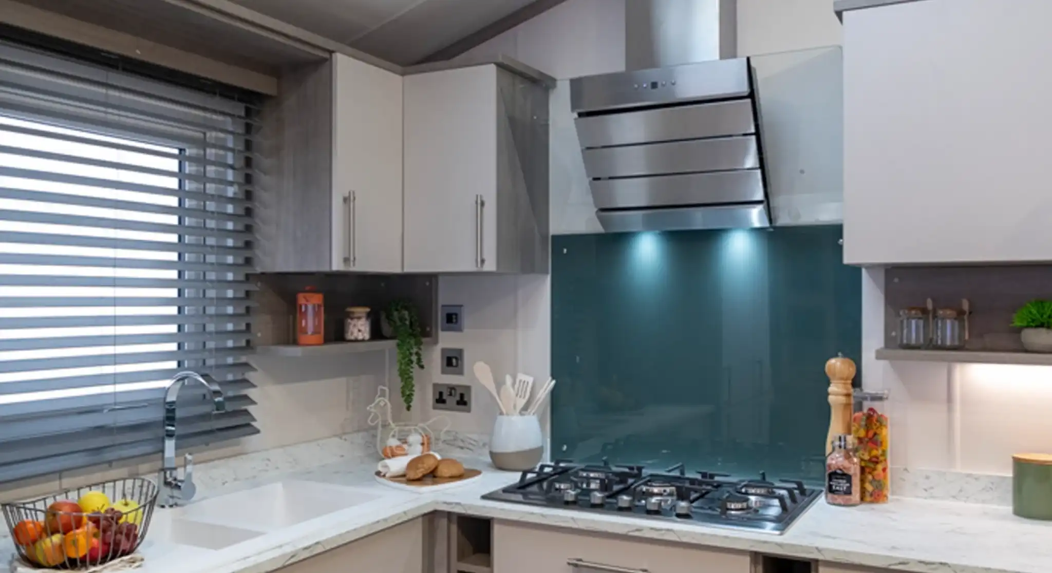 Contemporary kitchen with light cabinetry, a white marble-style countertop, and a teal glass backsplash behind a stainless steel gas cooktop and extractor hood. The kitchen includes a built-in sink below a window with modern horizontal blinds, open shelving with decorative items, and various kitchen essentials like utensils, a fruit basket, and jars