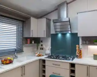 Contemporary kitchen with light cabinetry, a white marble-style countertop, and a teal glass backsplash behind a stainless steel gas cooktop and extractor hood. The kitchen includes a built-in sink below a window with modern horizontal blinds, open shelving with decorative items, and various kitchen essentials like utensils, a fruit basket, and jars