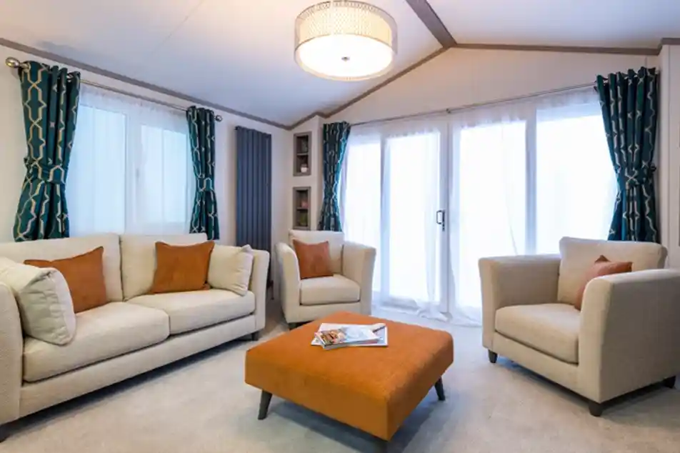 Luxurious lounge with beige sofas and armchairs decorated with rust-coloured pillows, a rust coloured oversized footstool that doubles up as a coffee table, and blue and gold patterned curtains either side of the windows and patio doors.