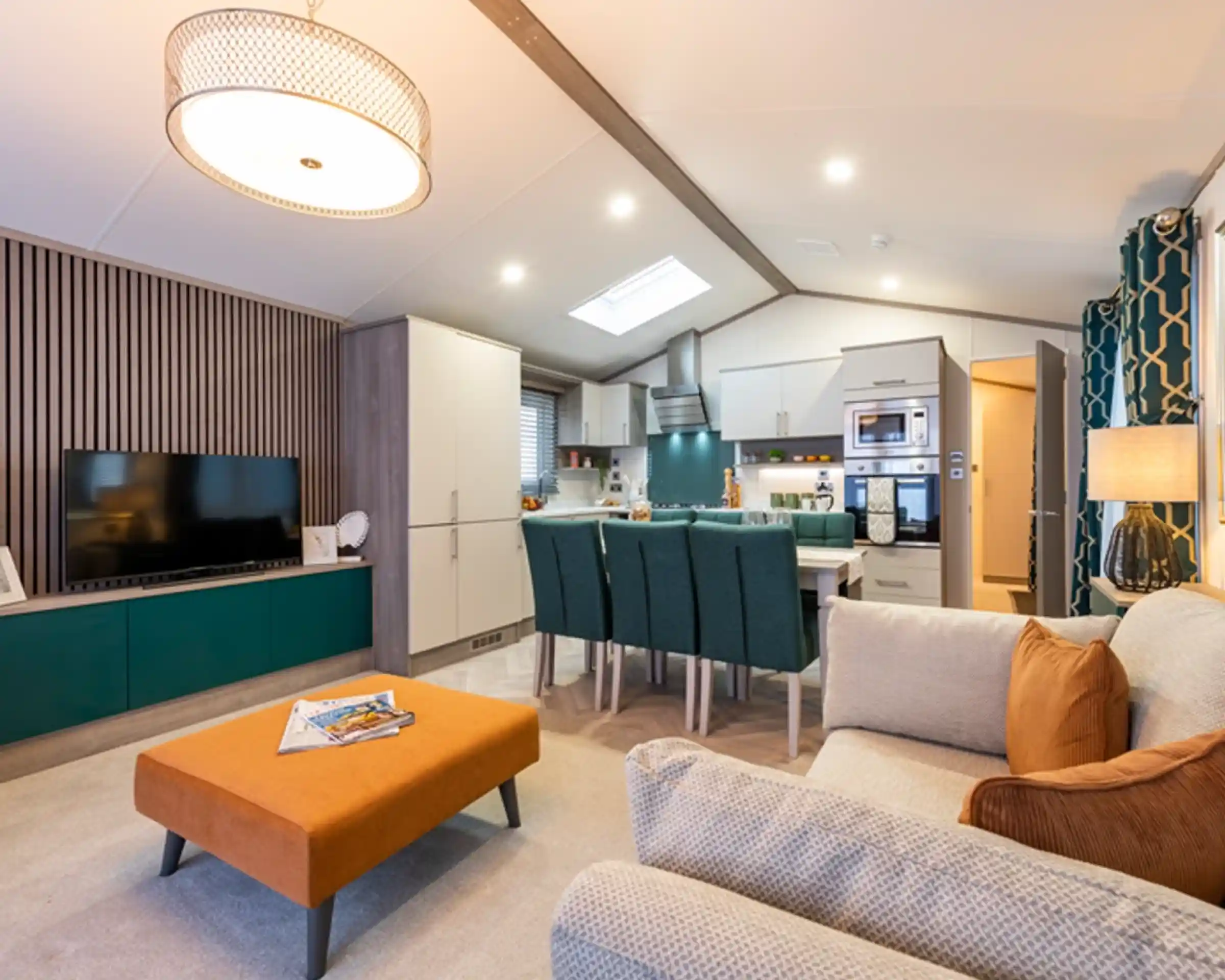 Modern open-plan living area with a kitchen, dining, and lounge space. The room features a cozy beige sofa with orange cushions, a matching orange ottoman, a flat-screen TV on a teal and wood entertainment unit, a dining table with teal chairs, and a fully equipped kitchen with integrated appliances. Contemporary lighting and decorative elements enhance the stylish and welcoming interior.