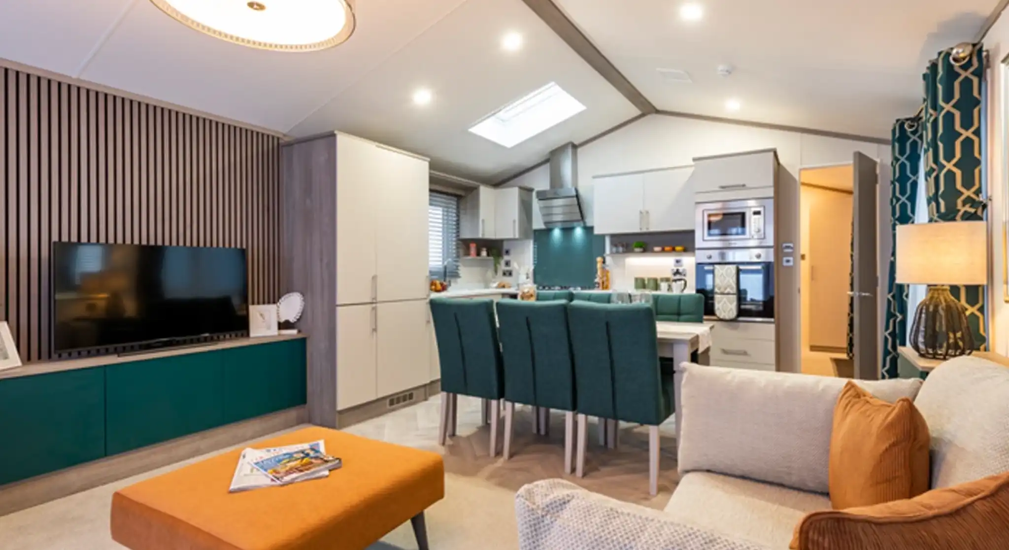 Modern open-plan living area with a kitchen, dining, and lounge space. The room features a cozy beige sofa with orange cushions, a matching orange ottoman, a flat-screen TV on a teal and wood entertainment unit, a dining table with teal chairs, and a fully equipped kitchen with integrated appliances. Contemporary lighting and decorative elements enhance the stylish and welcoming interior.