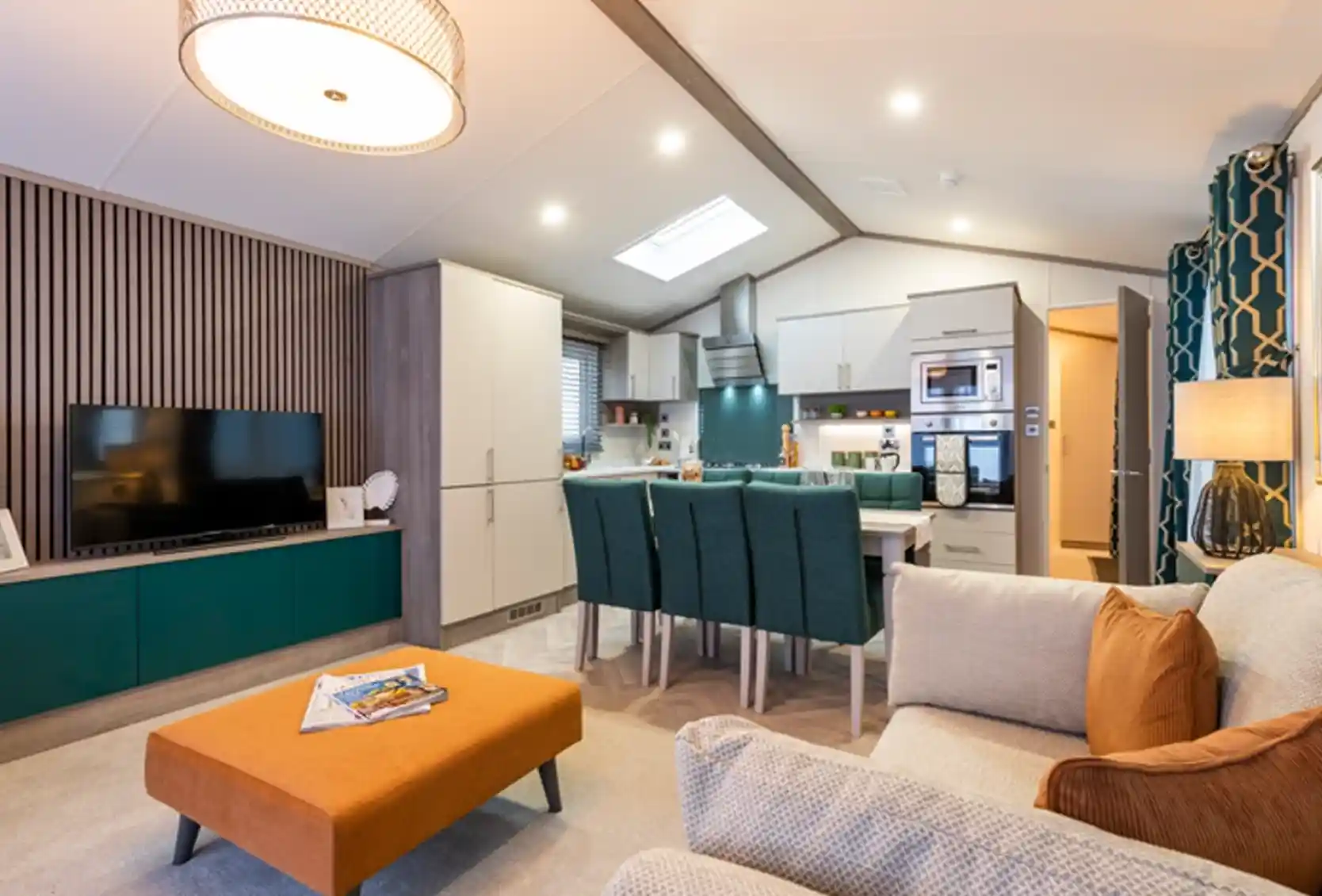 Modern open-plan living area with a kitchen, dining, and lounge space. The room features a cozy beige sofa with orange cushions, a matching orange ottoman, a flat-screen TV on a teal and wood entertainment unit, a dining table with teal chairs, and a fully equipped kitchen with integrated appliances. Contemporary lighting and decorative elements enhance the stylish and welcoming interior.