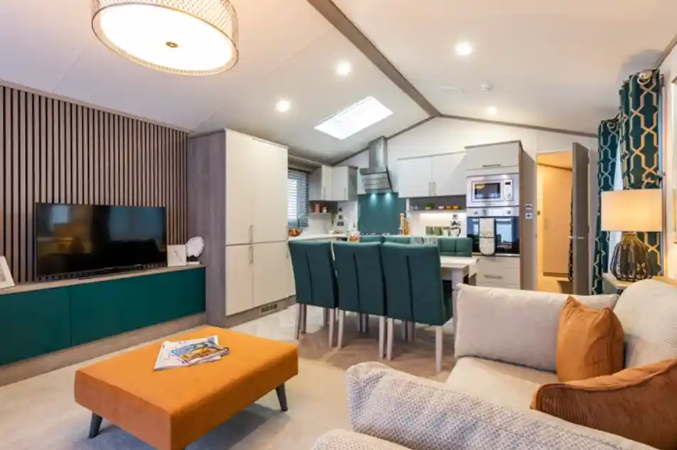 Modern open-plan living area with a kitchen, dining, and lounge space. The room features a cozy beige sofa with orange cushions, a matching orange ottoman, a flat-screen TV on a teal and wood entertainment unit, a dining table with teal chairs, and a fully equipped kitchen with integrated appliances. Contemporary lighting and decorative elements enhance the stylish and welcoming interior.