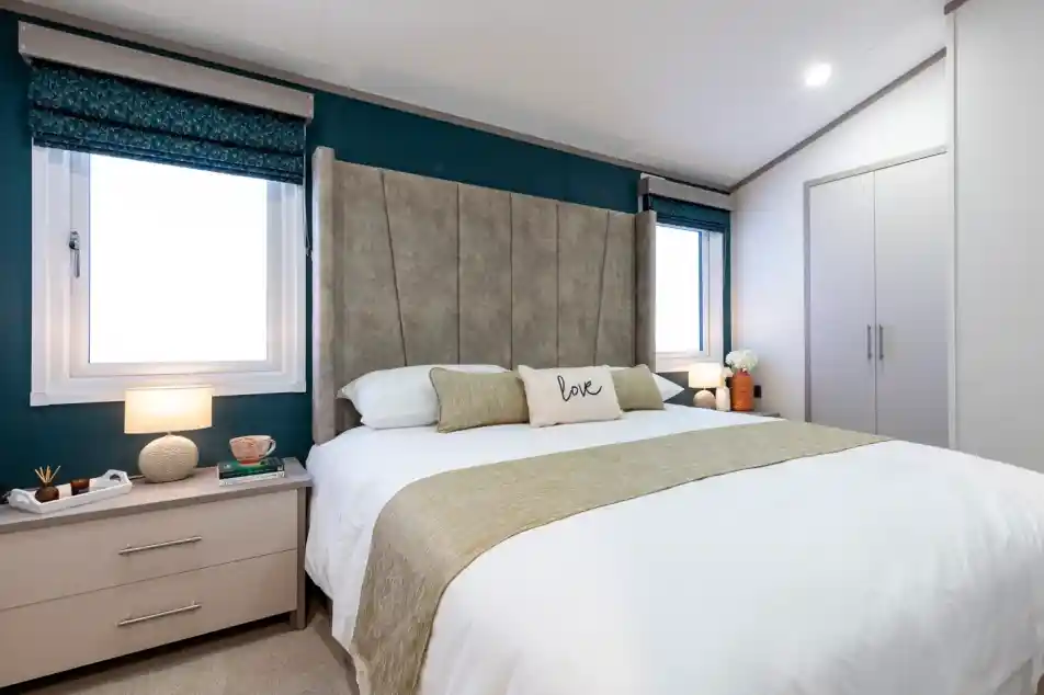 Elegant master bedroom with a large upholstered headboard, a double bed dressed in white linens and gold cushions and throw. The room features two windows with patterned Roman blinds, bedside tables with lamps and decorative items, and a built-in wardrobe with sleek doors.