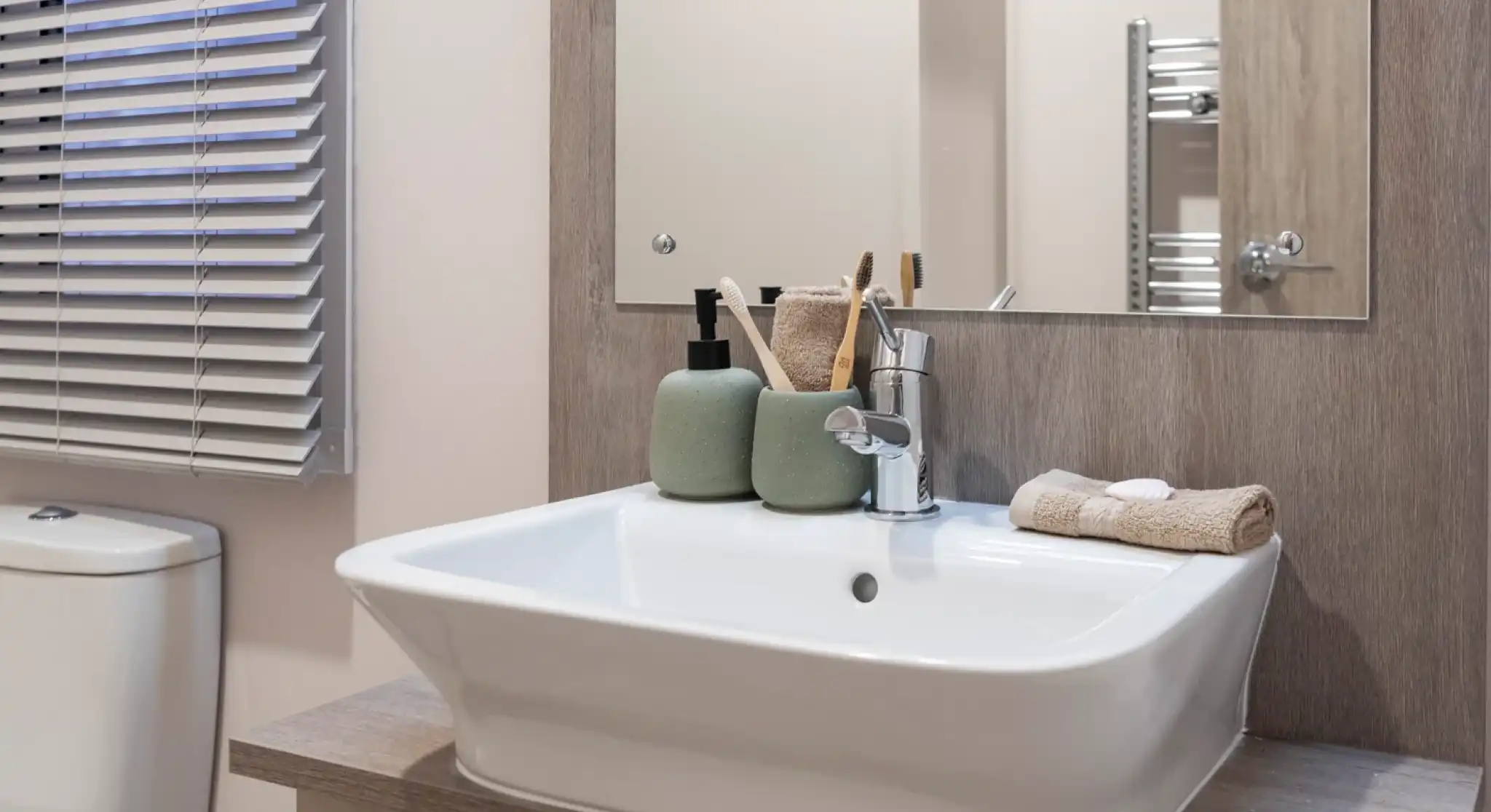 Modern bathroom vanity area featuring a rectangular white basin with a chrome mixer tap, set on a wood-effect countertop. Above the sink is a large mirror with a wooden frame. Accessories include green ceramic soap dispensers, toothbrush holders, and neatly folded beige towels. A toilet and window with horizontal blinds are partially visible.