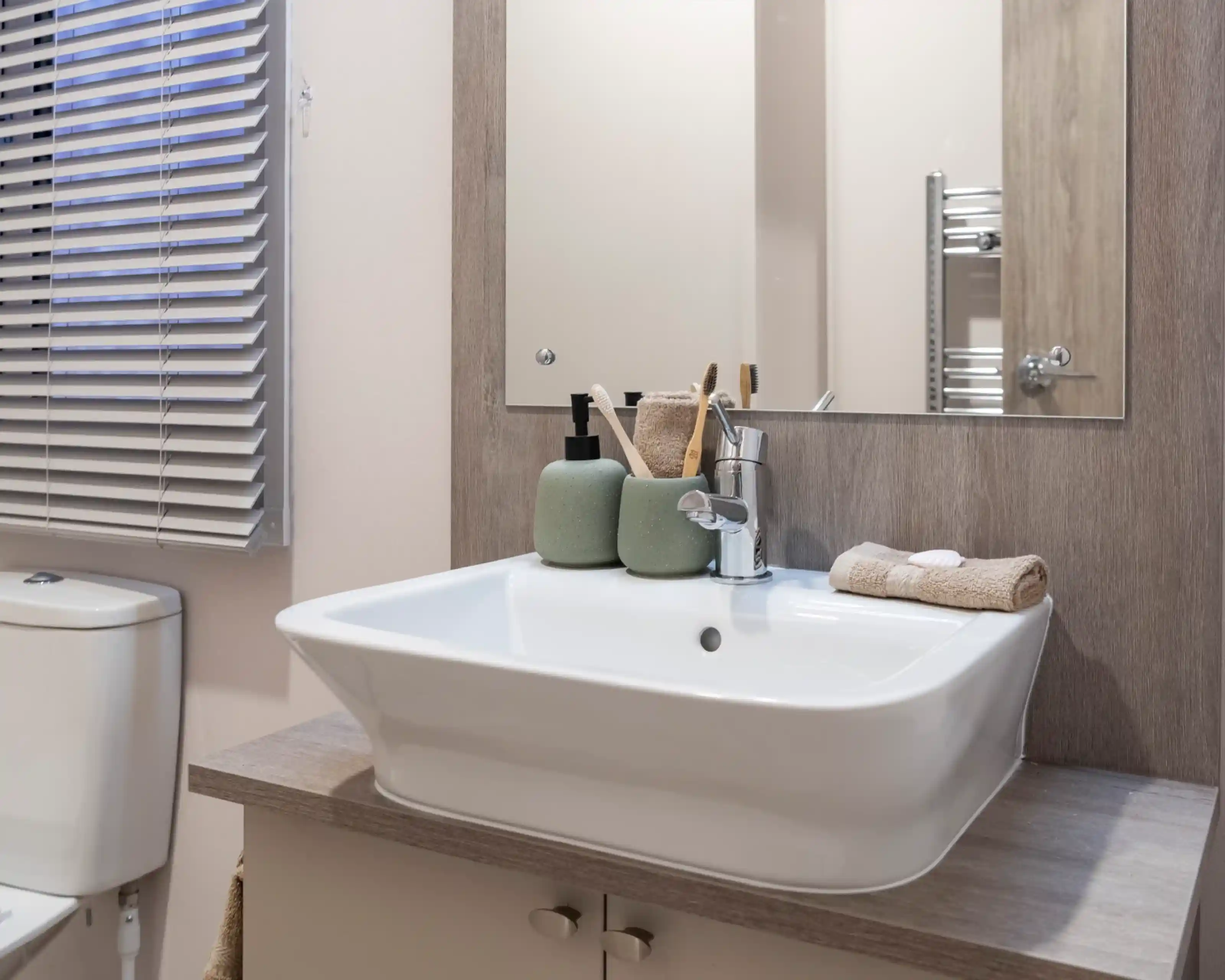 Modern bathroom vanity area featuring a rectangular white basin with a chrome mixer tap, set on a wood-effect countertop. Above the sink is a large mirror with a wooden frame. Accessories include green ceramic soap dispensers, toothbrush holders, and neatly folded beige towels. A toilet and window with horizontal blinds are partially visible.