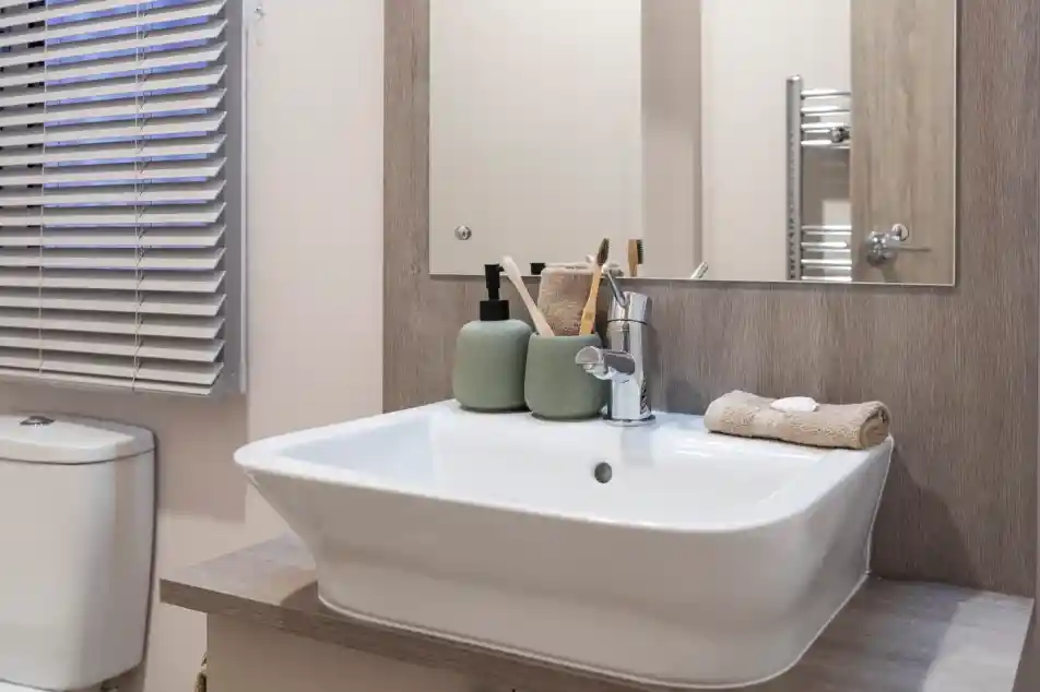 Modern bathroom vanity area featuring a rectangular white basin with a chrome mixer tap, set on a wood-effect countertop. Above the sink is a large mirror with a wooden frame. Accessories include green ceramic soap dispensers, toothbrush holders, and neatly folded beige towels. A toilet and window with horizontal blinds are partially visible.