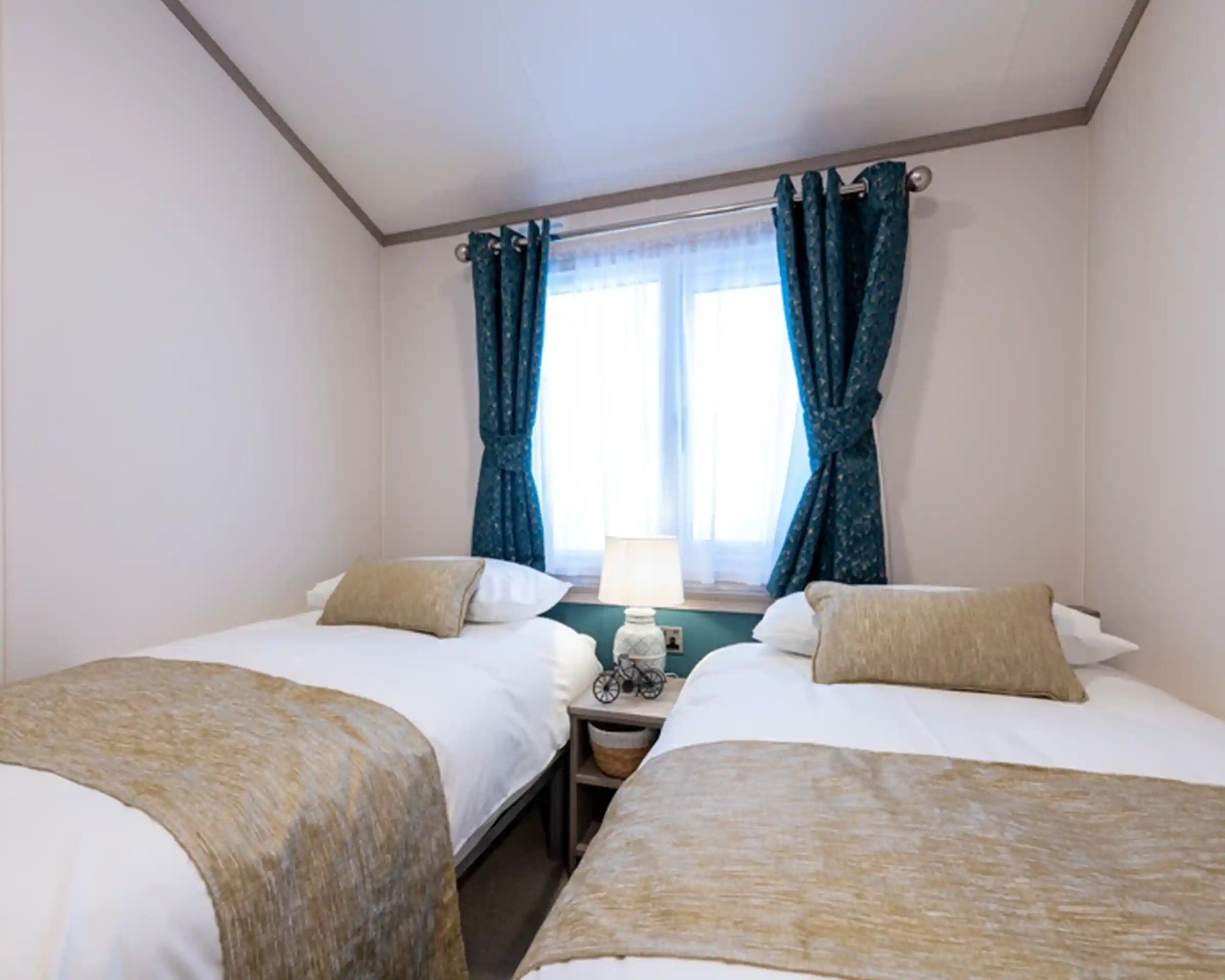 Cozy twin bedroom with two single beds dressed in white linens and beige throws and cushions. A small nightstand with a decorative lamp sits between the beds beneath a window with sheer white curtains and teal patterned drapes.