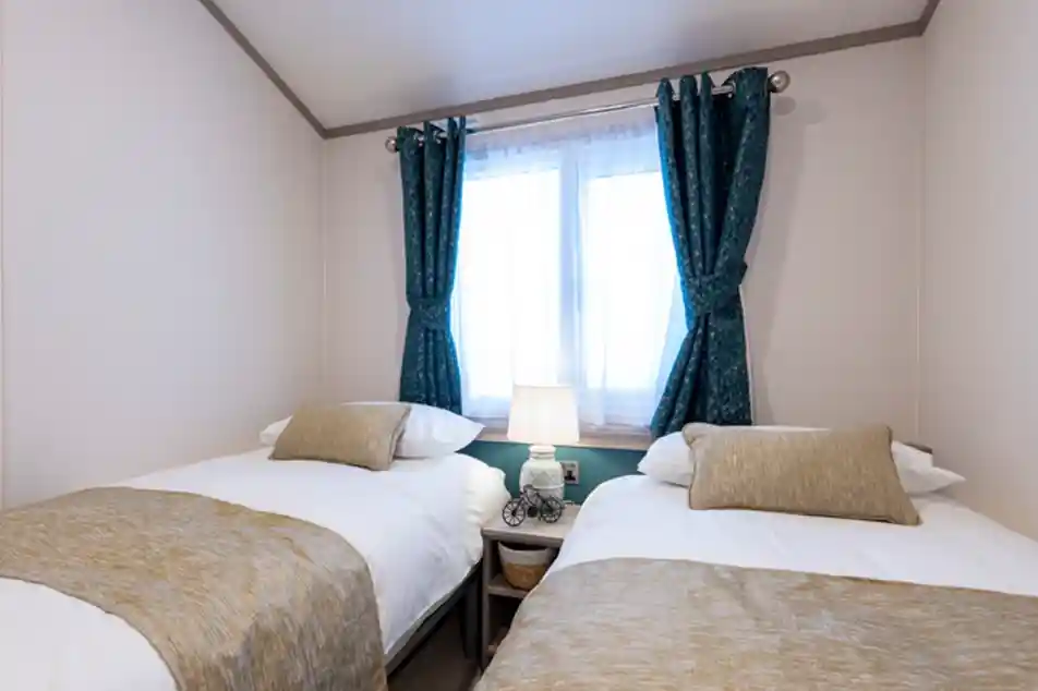 Cozy twin bedroom with two single beds dressed in white linens and beige throws and cushions. A small nightstand with a decorative lamp sits between the beds beneath a window with sheer white curtains and teal patterned drapes.