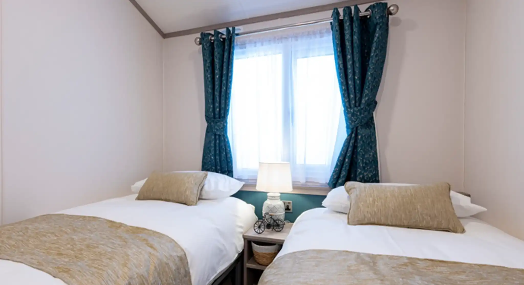 Cozy twin bedroom with two single beds dressed in white linens and beige throws and cushions. A small nightstand with a decorative lamp sits between the beds beneath a window with sheer white curtains and teal patterned drapes.