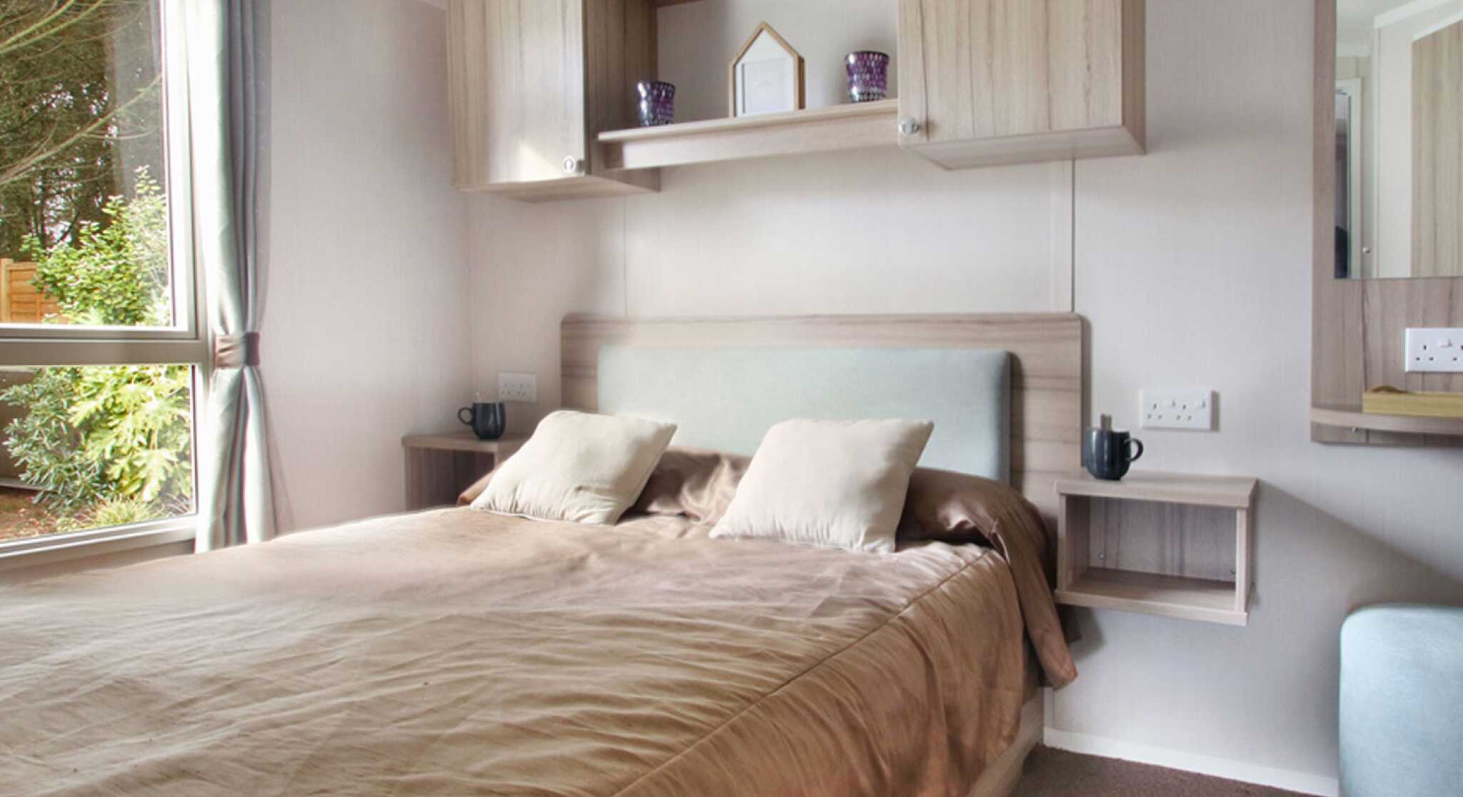 Modern bedroom with a cozy brown bed, light wood furniture, and two bedside shelves. A large window allows natural light to brighten the space.