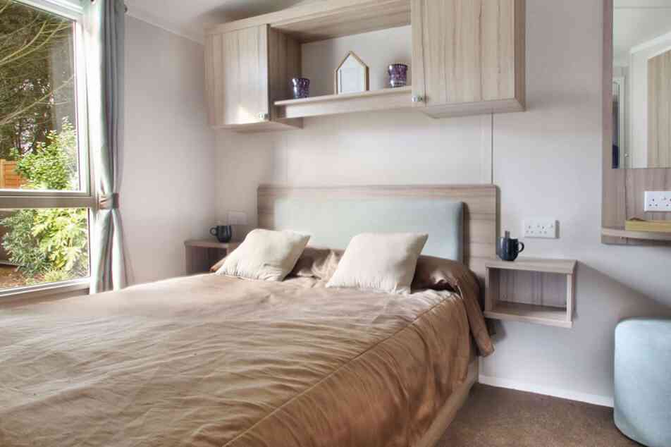 Modern bedroom with a cozy brown bed, light wood furniture, and two bedside shelves. A large window allows natural light to brighten the space.