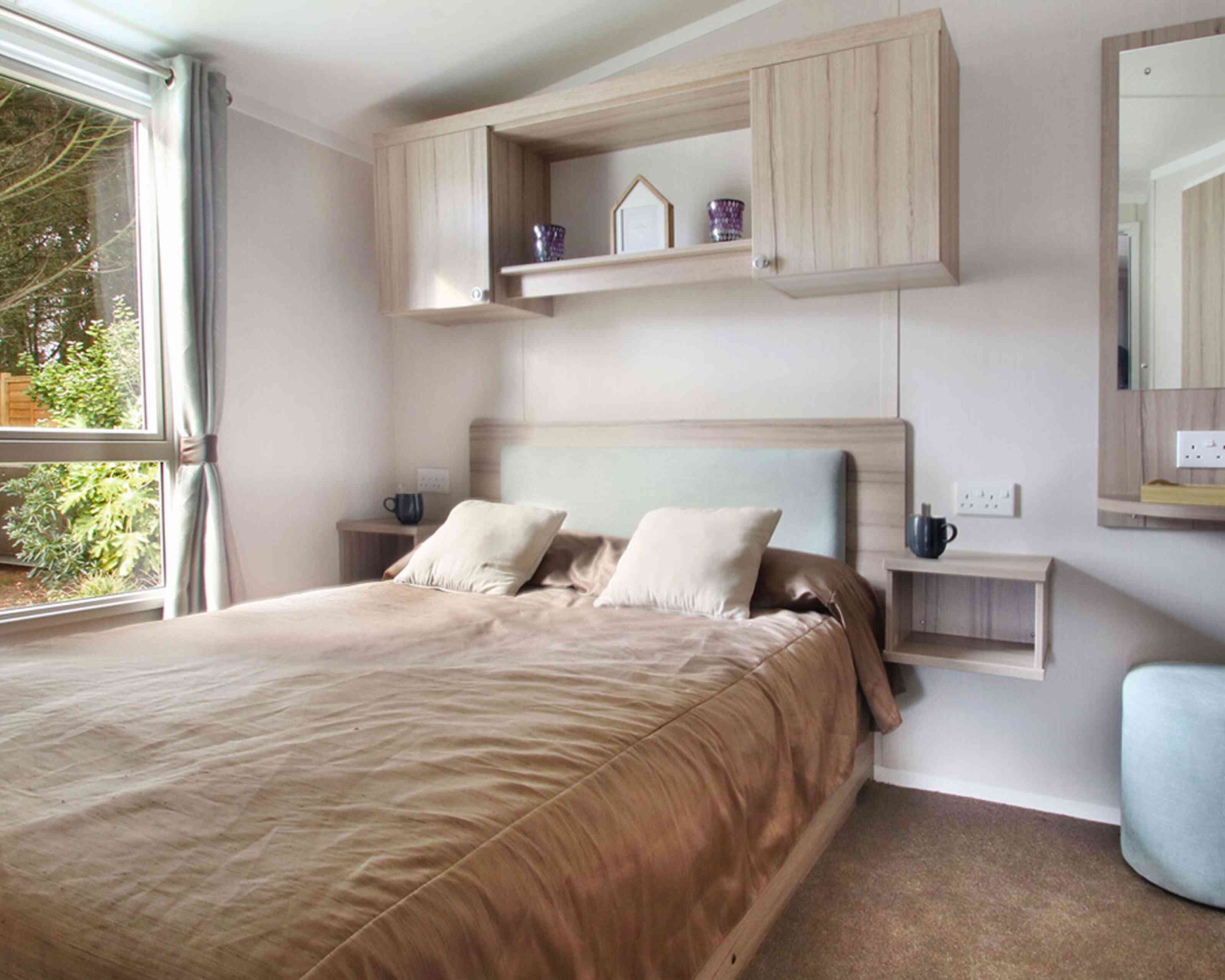 Modern bedroom with a cozy brown bed, light wood furniture, and two bedside shelves. A large window allows natural light to brighten the space.