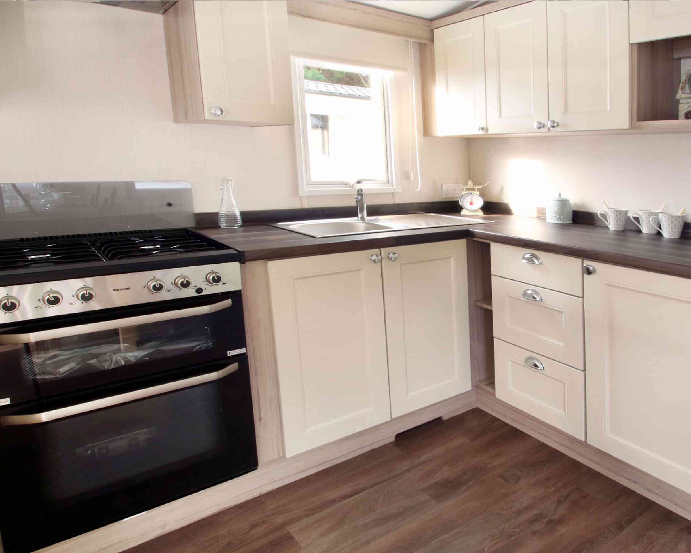 Bright, modern kitchen and living area in neutral tones with wood accents. Dining table set for two, floral curtains, and cozy seating enhance warmth.