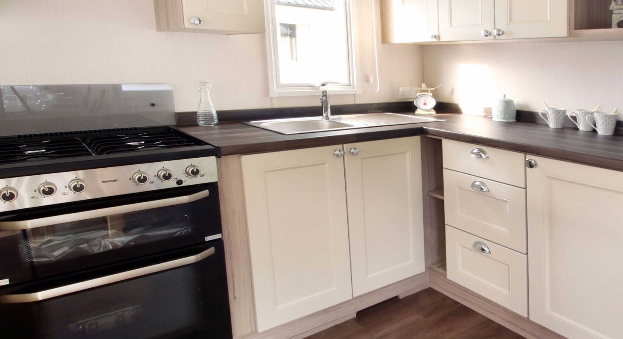Bright, modern kitchen and living area in neutral tones with wood accents. Dining table set for two, floral curtains, and cozy seating enhance warmth.