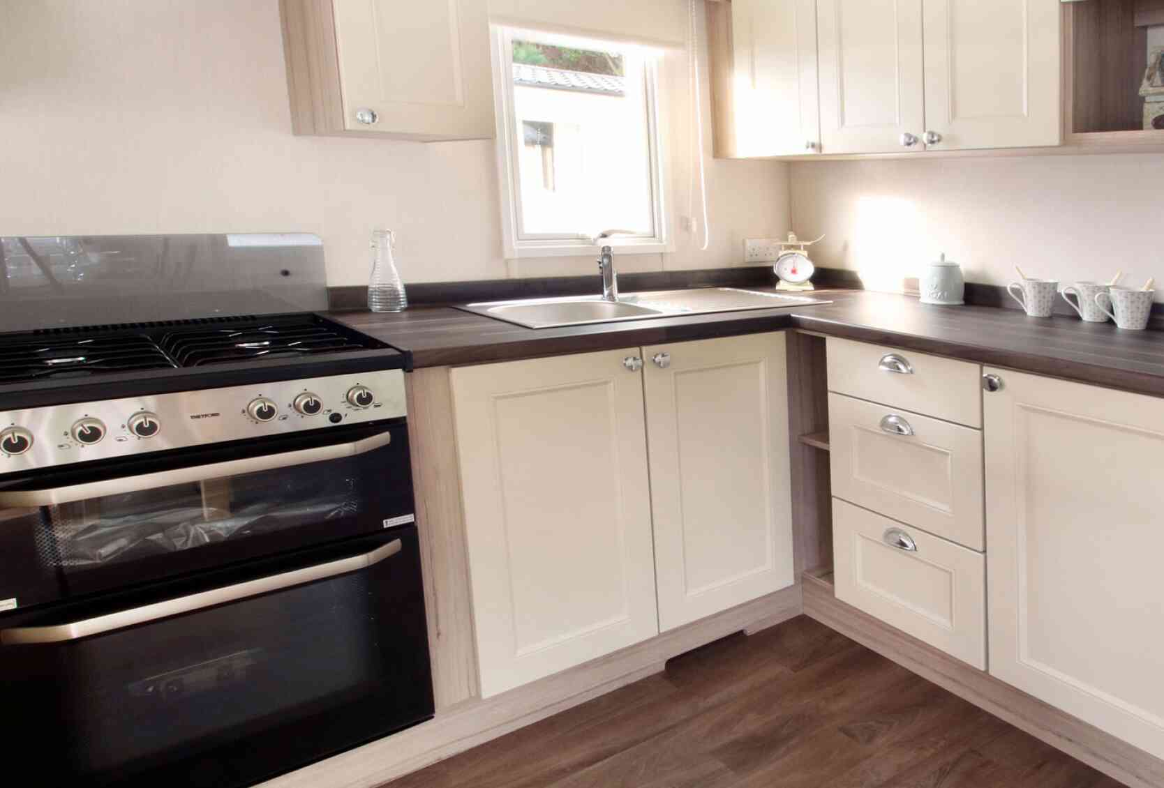 Bright, modern kitchen and living area in neutral tones with wood accents. Dining table set for two, floral curtains, and cozy seating enhance warmth.