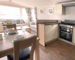 A cozy, well-lit living area with a beige sofa, floral curtains, and a small kitchen. A dining table set with plates and glasses is in the foreground.