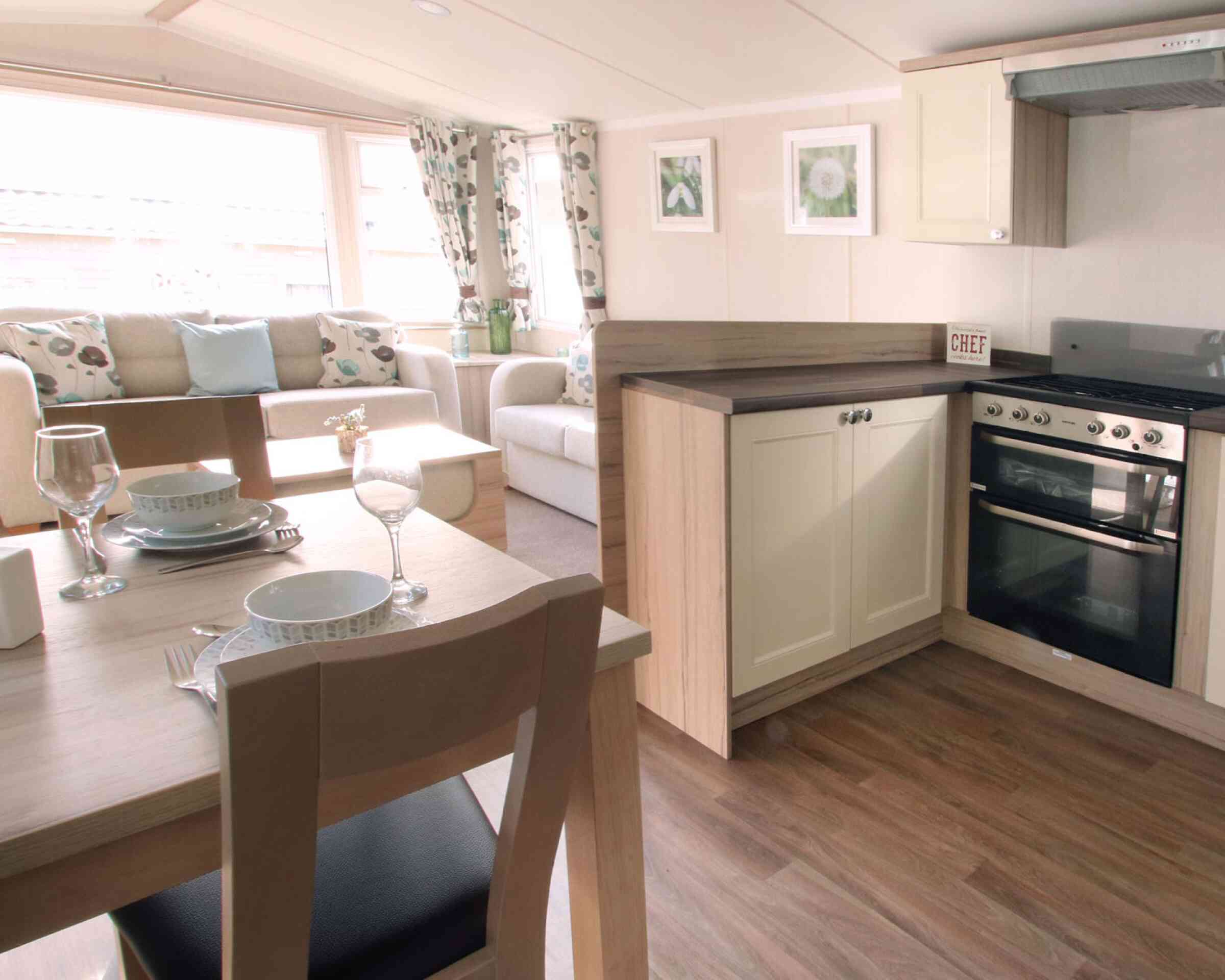 A cozy, well-lit living area with a beige sofa, floral curtains, and a small kitchen. A dining table set with plates and glasses is in the foreground.