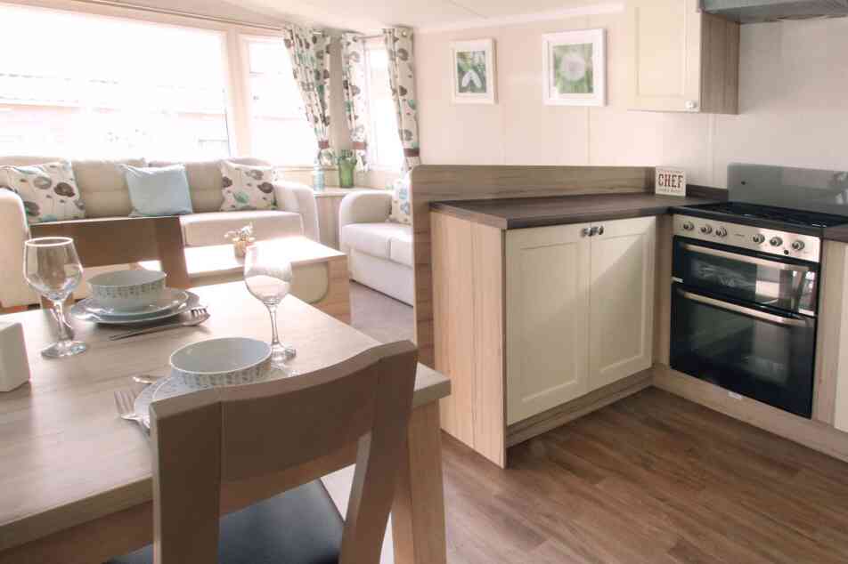 A cozy, well-lit living area with a beige sofa, floral curtains, and a small kitchen. A dining table set with plates and glasses is in the foreground.
