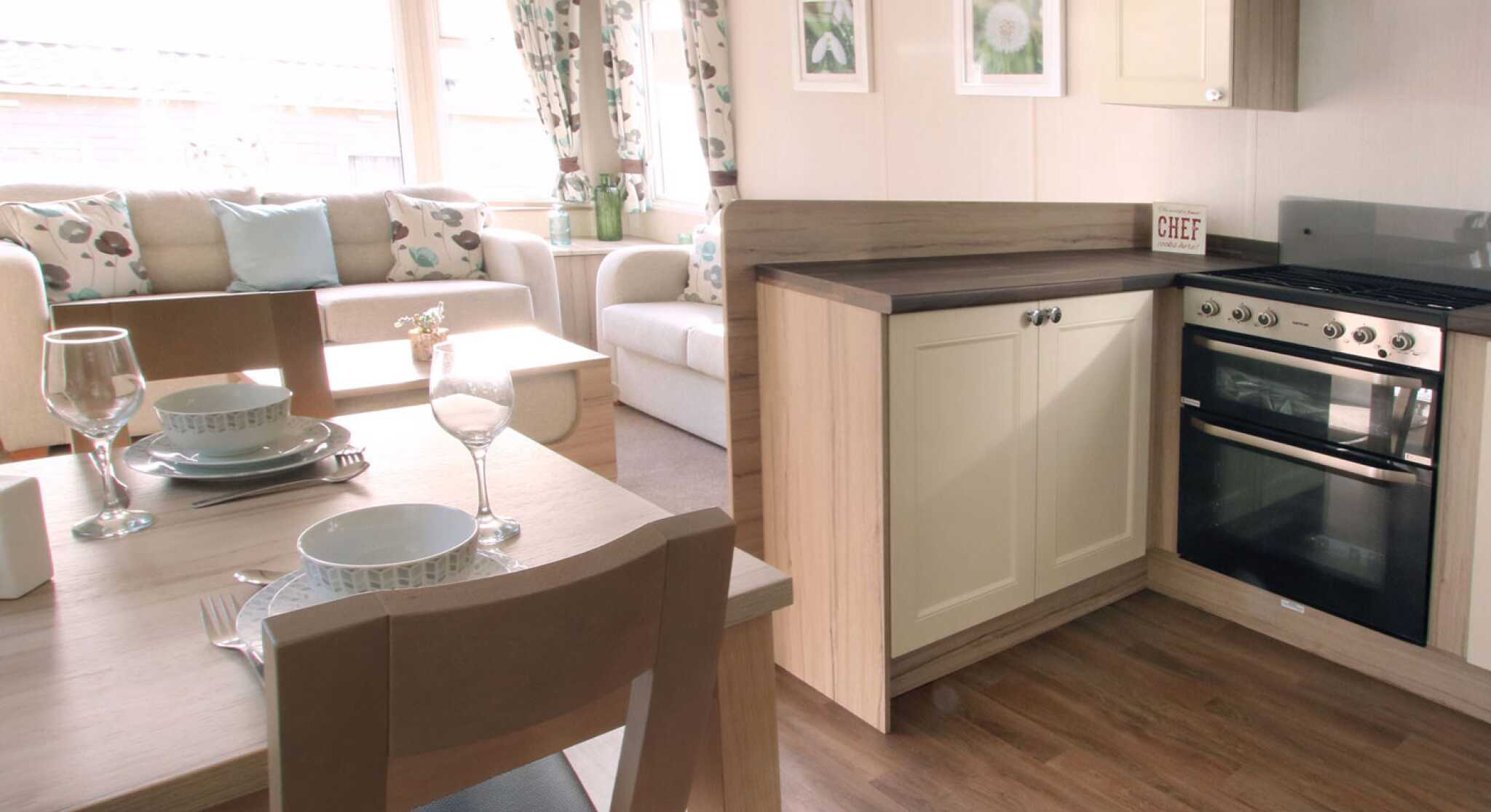 A cozy, well-lit living area with a beige sofa, floral curtains, and a small kitchen. A dining table set with plates and glasses is in the foreground.