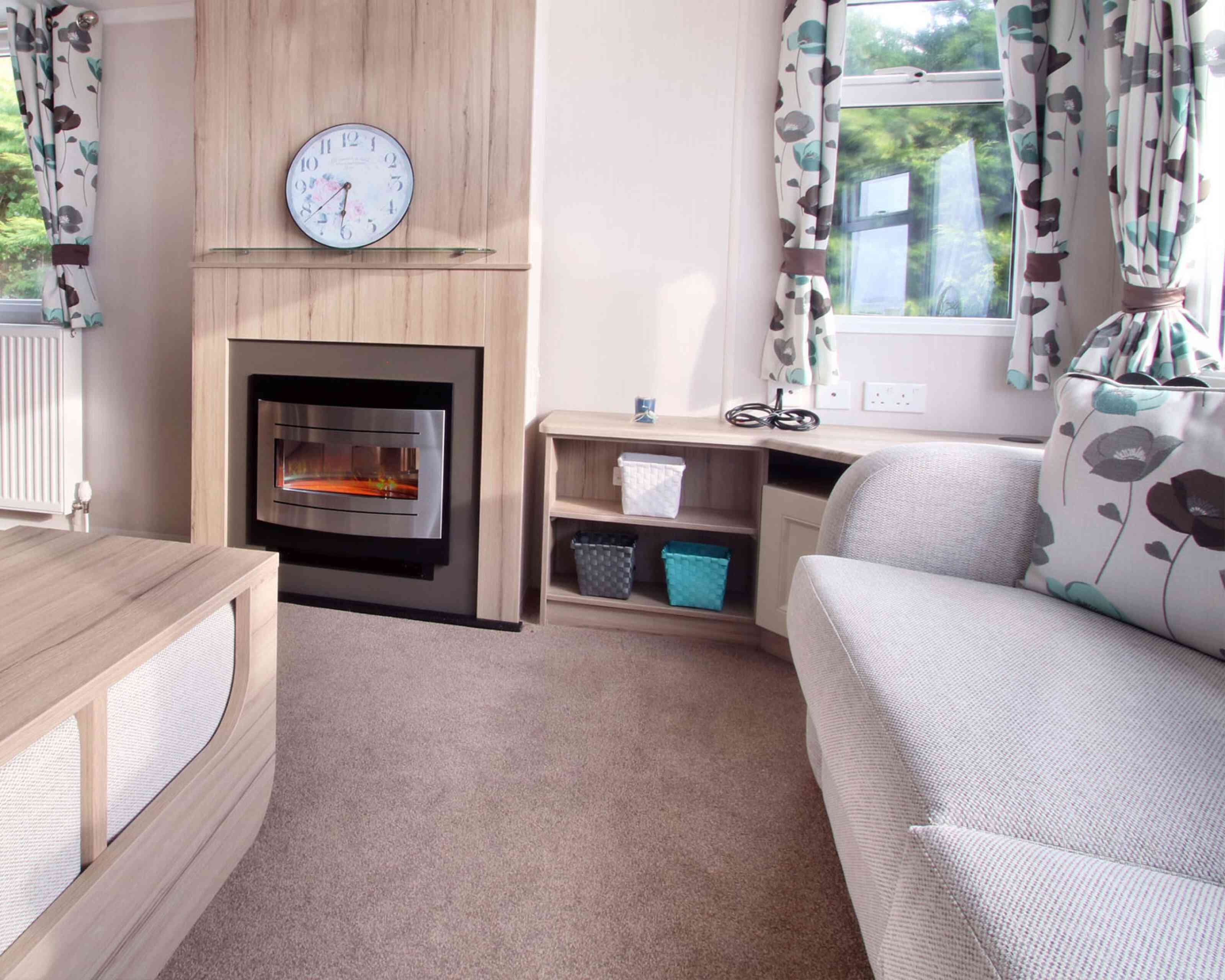 Cozy living room with a grey couch, floral curtains, electric fireplace, and clock above. Shelves hold woven baskets; sunlight filters through windows.