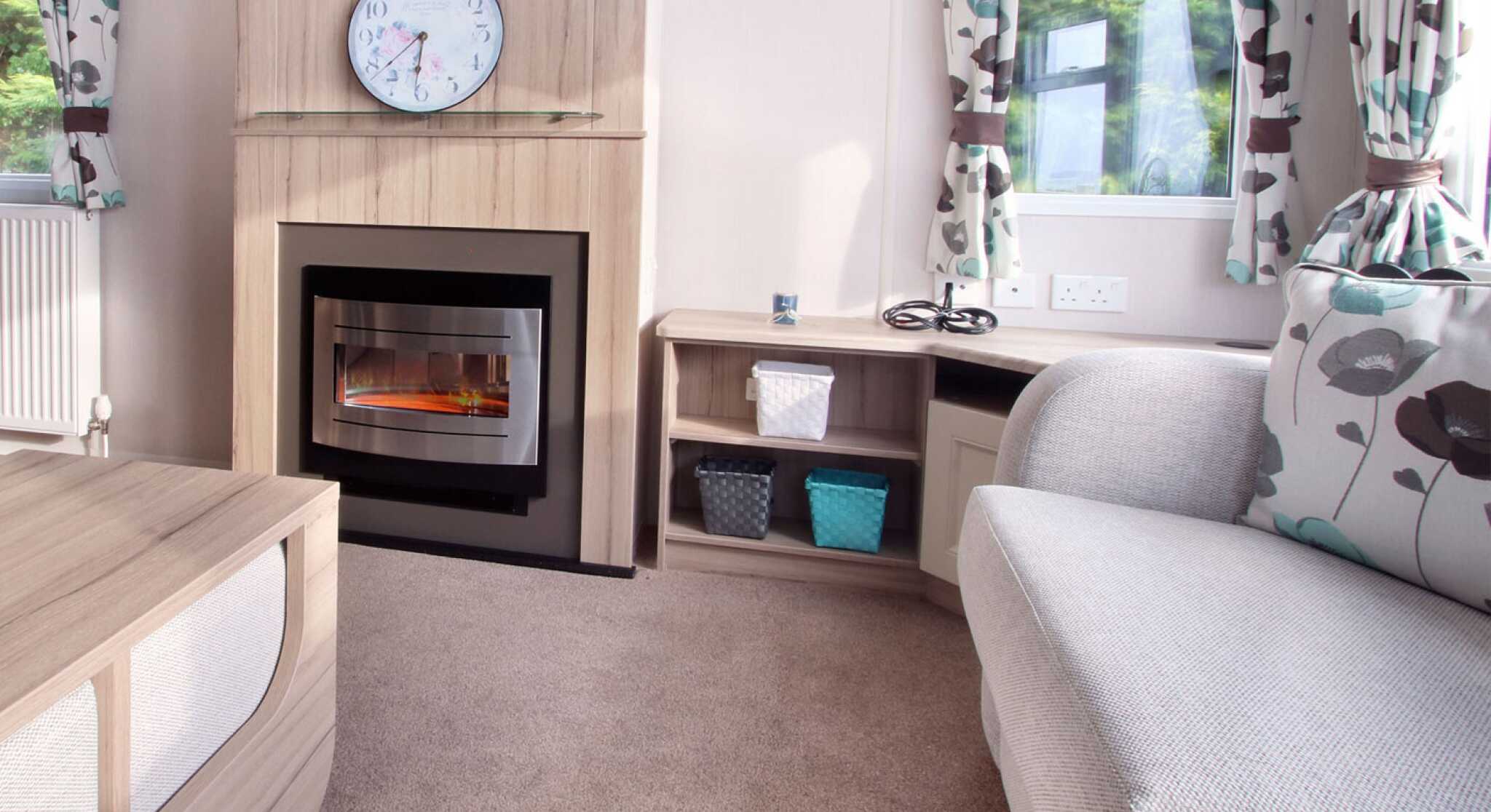 Cozy living room with a grey couch, floral curtains, electric fireplace, and clock above. Shelves hold woven baskets; sunlight filters through windows.