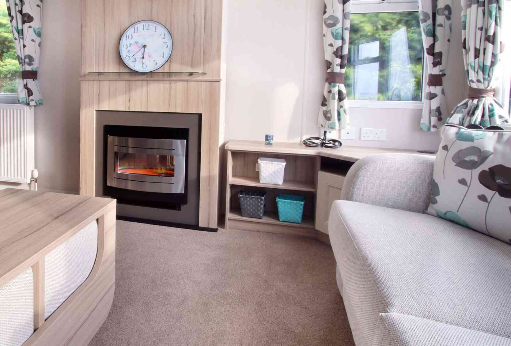 Cozy living room with a grey couch, floral curtains, electric fireplace, and clock above. Shelves hold woven baskets; sunlight filters through windows.