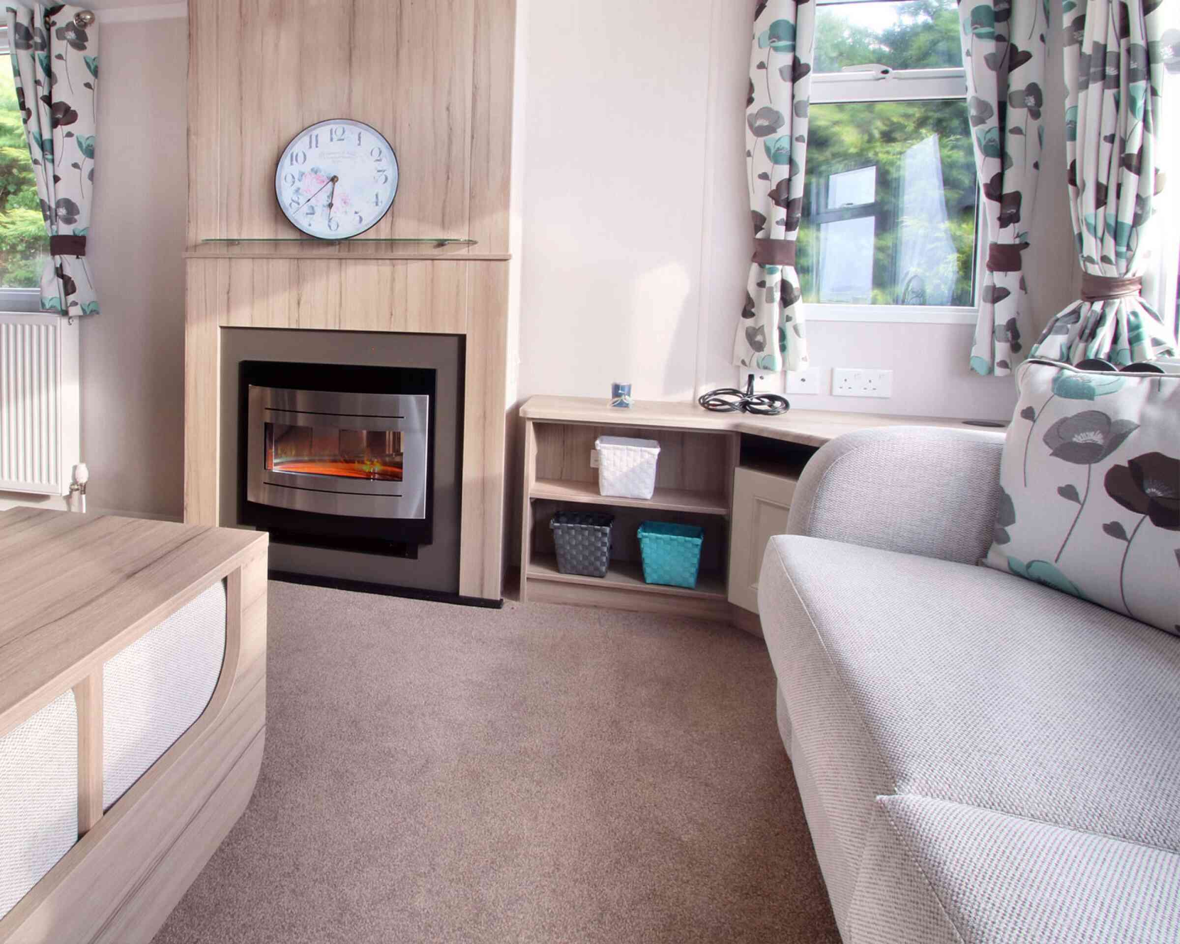 Cozy living room with a grey couch, floral curtains, electric fireplace, and clock above. Shelves hold woven baskets; sunlight filters through windows.