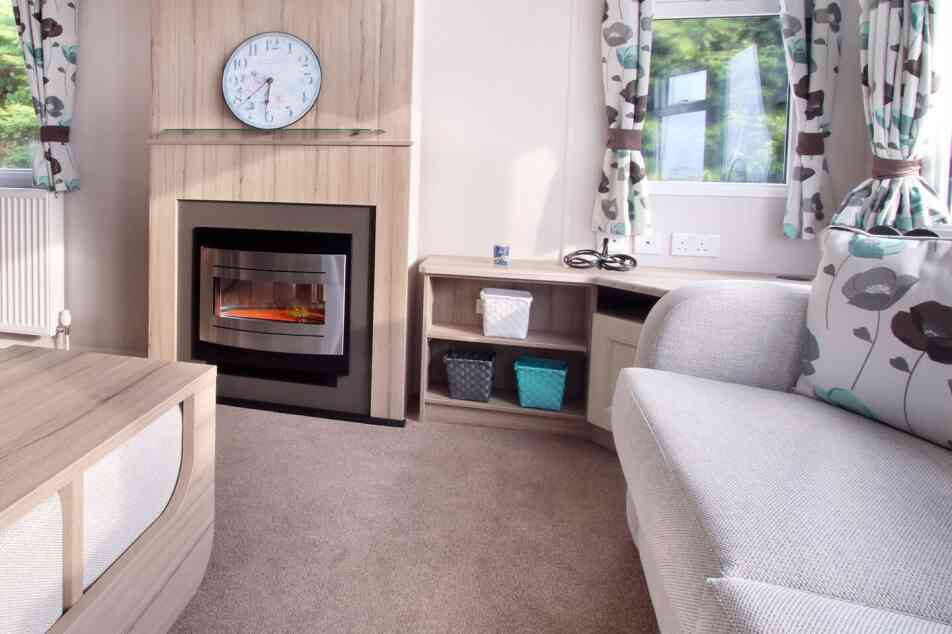 Cozy living room with a grey couch, floral curtains, electric fireplace, and clock above. Shelves hold woven baskets; sunlight filters through windows.