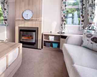 Cozy living room with a grey couch, floral curtains, electric fireplace, and clock above. Shelves hold woven baskets; sunlight filters through windows.