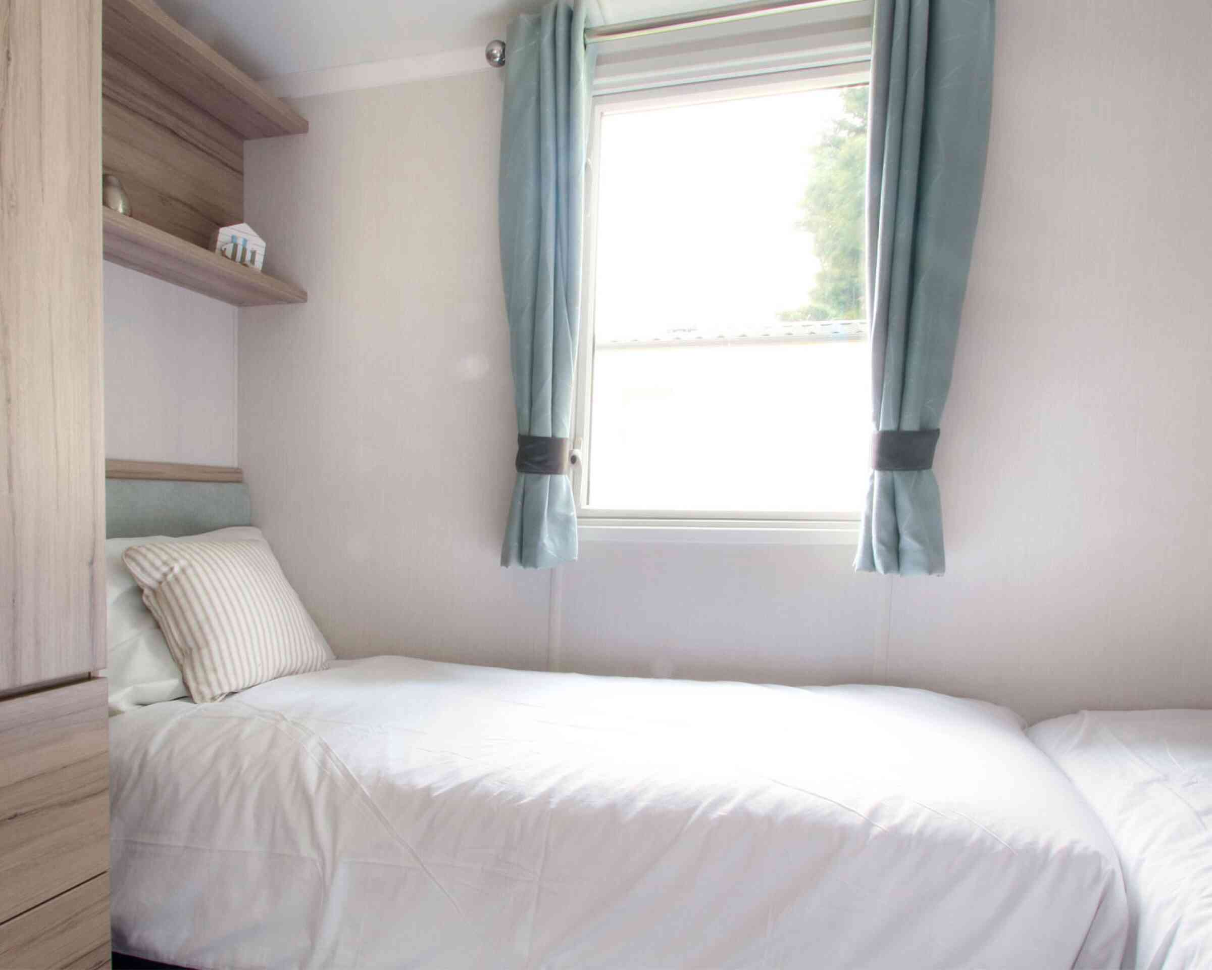 Small, bright bedroom with two single beds, white bedding, and a striped pillows. Light blue curtains frame a window. Wooden shelves and a minimalist decor.