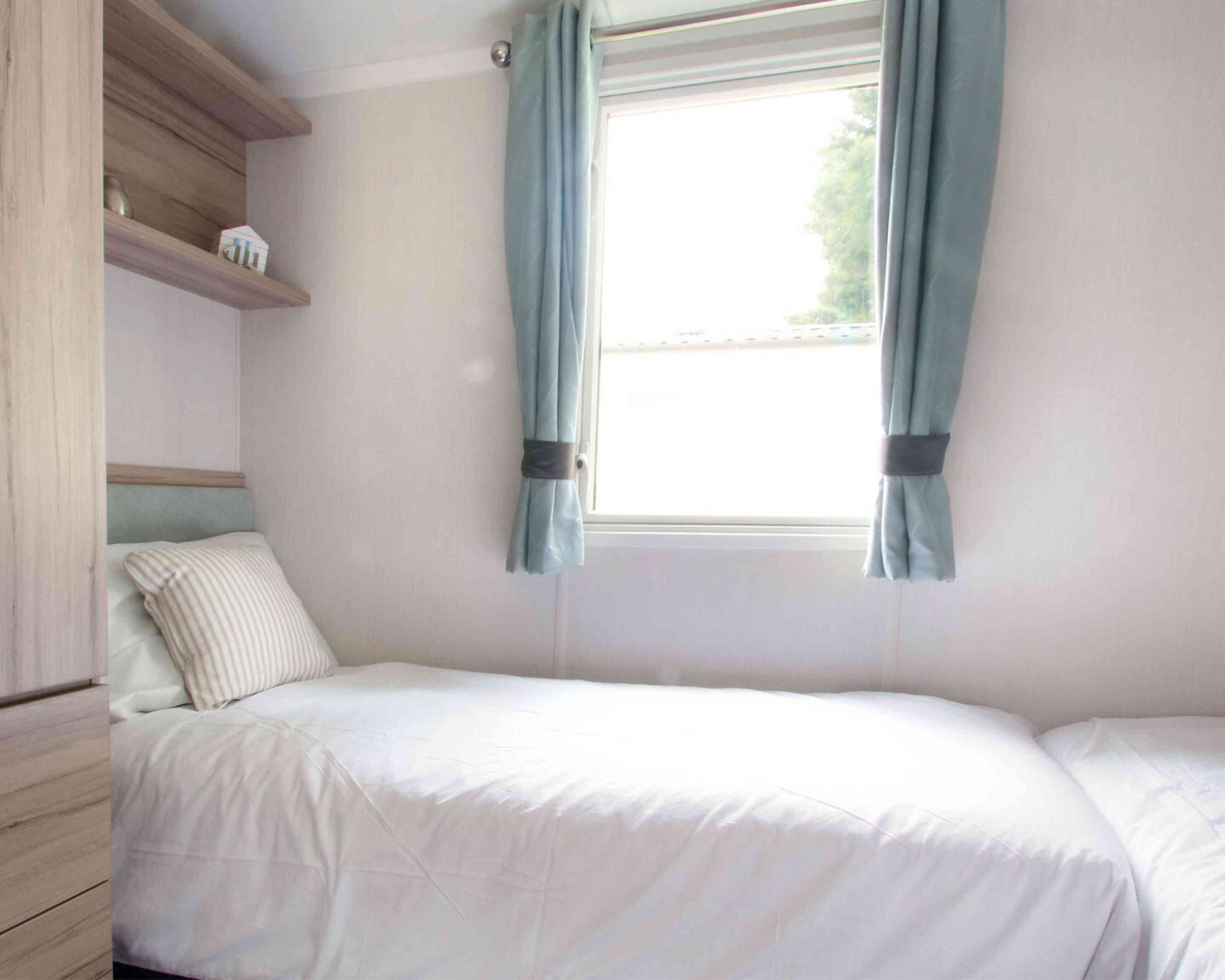 Small, bright bedroom with two single beds, white bedding, and a striped pillows. Light blue curtains frame a window. Wooden shelves and a minimalist decor.