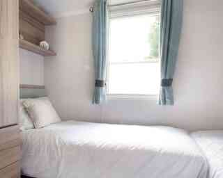 Small, bright bedroom with two single beds, white bedding, and a striped pillows. Light blue curtains frame a window. Wooden shelves and a minimalist decor.