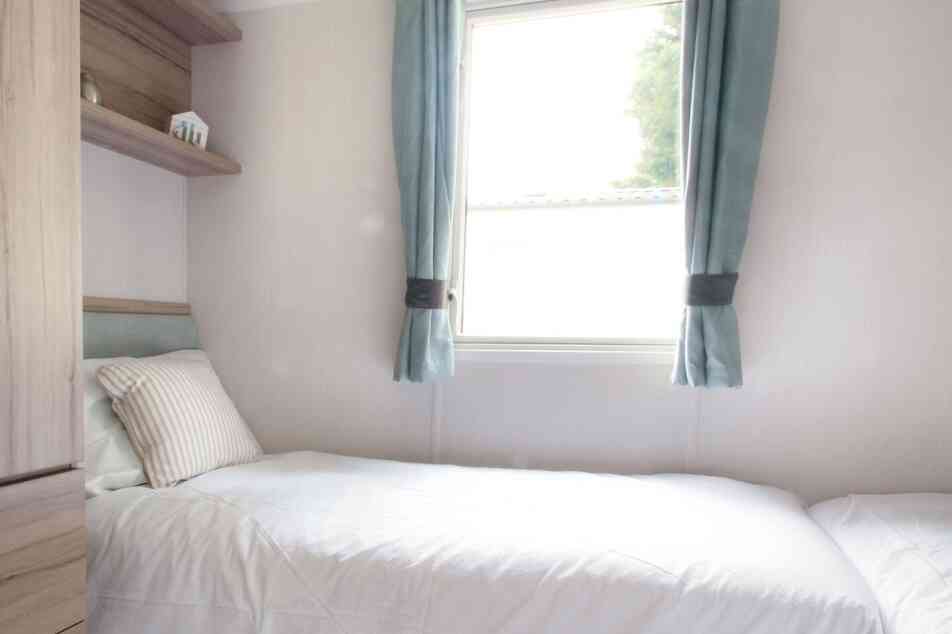 Small, bright bedroom with two single beds, white bedding, and a striped pillows. Light blue curtains frame a window. Wooden shelves and a minimalist decor.