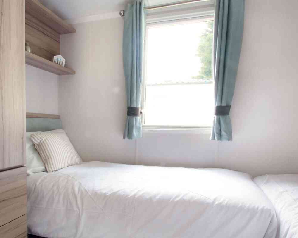 Small, bright bedroom with two single beds, white bedding, and a striped pillows. Light blue curtains frame a window. Wooden shelves and a minimalist decor.