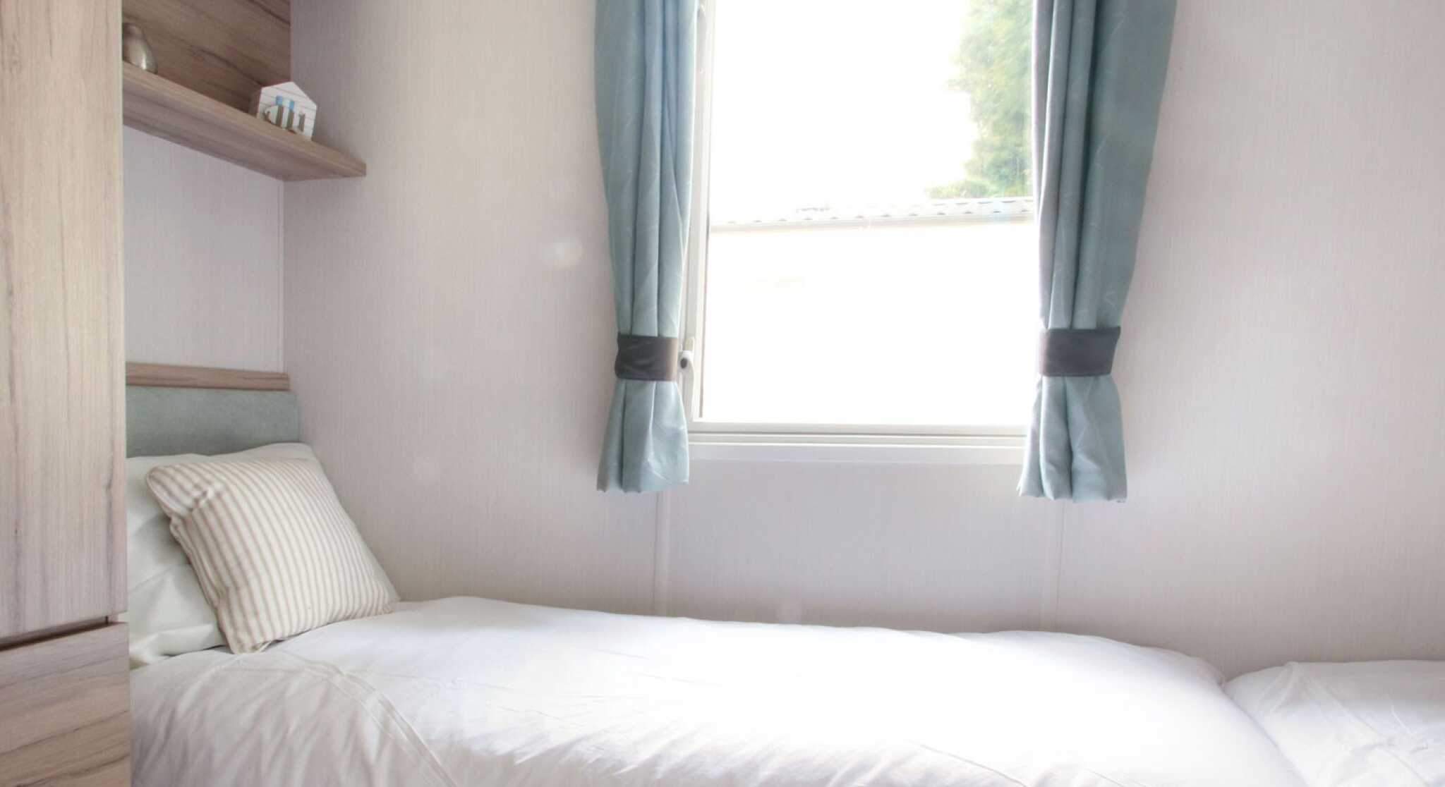 Small, bright bedroom with two single beds, white bedding, and a striped pillows. Light blue curtains frame a window. Wooden shelves and a minimalist decor.