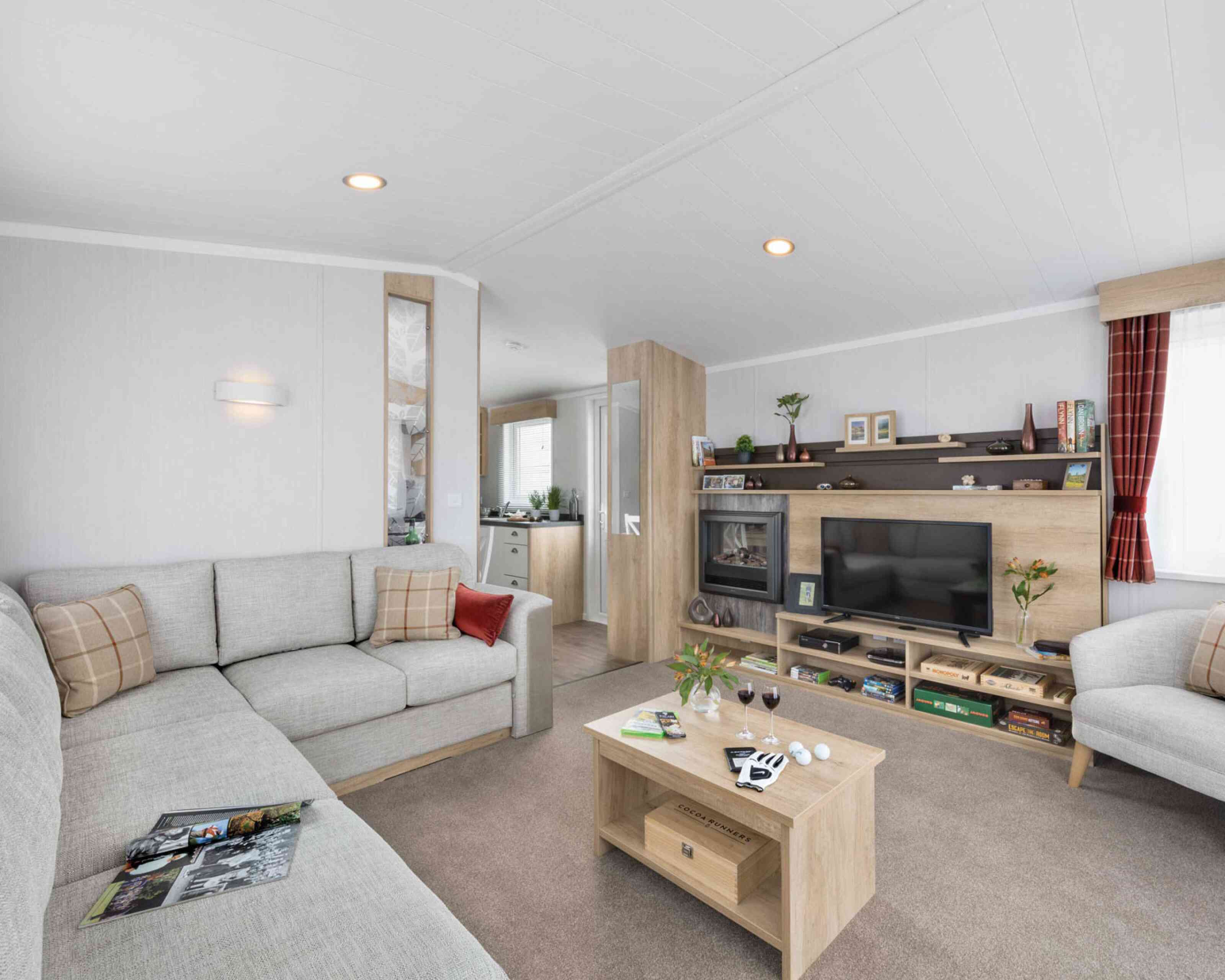 Inviting contemporary lounge featuring a grey corner sofa with patterned cushions, coffee table, TV, shelves, and soft, welcoming accents.
