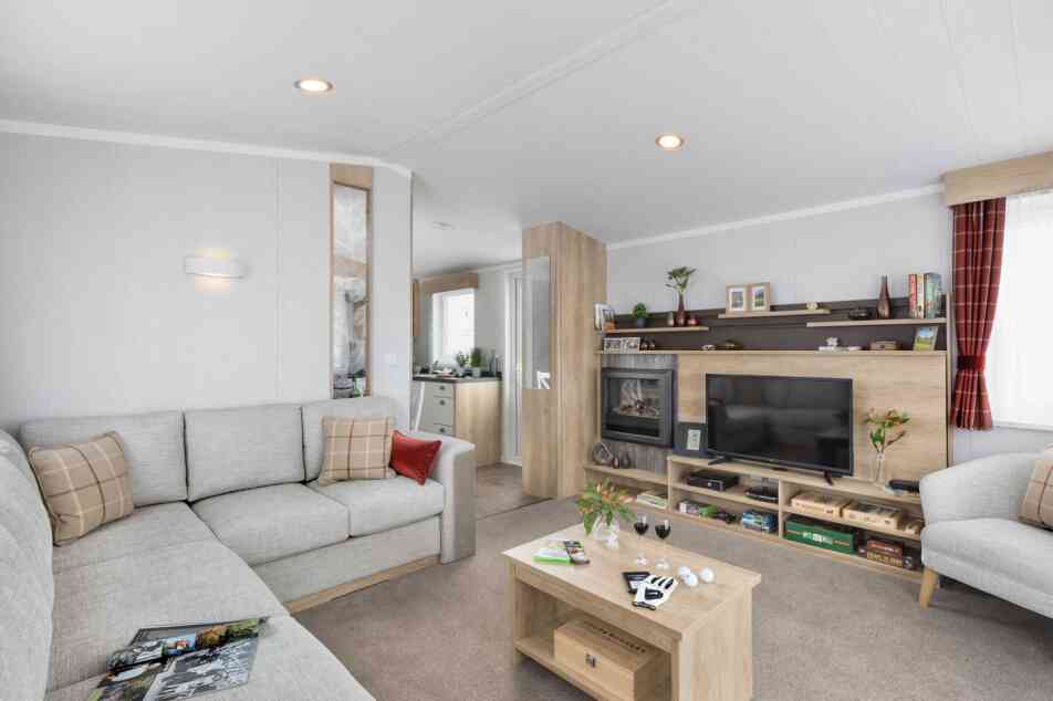 Inviting contemporary lounge featuring a grey corner sofa with patterned cushions, coffee table, TV, shelves, and soft, welcoming accents.