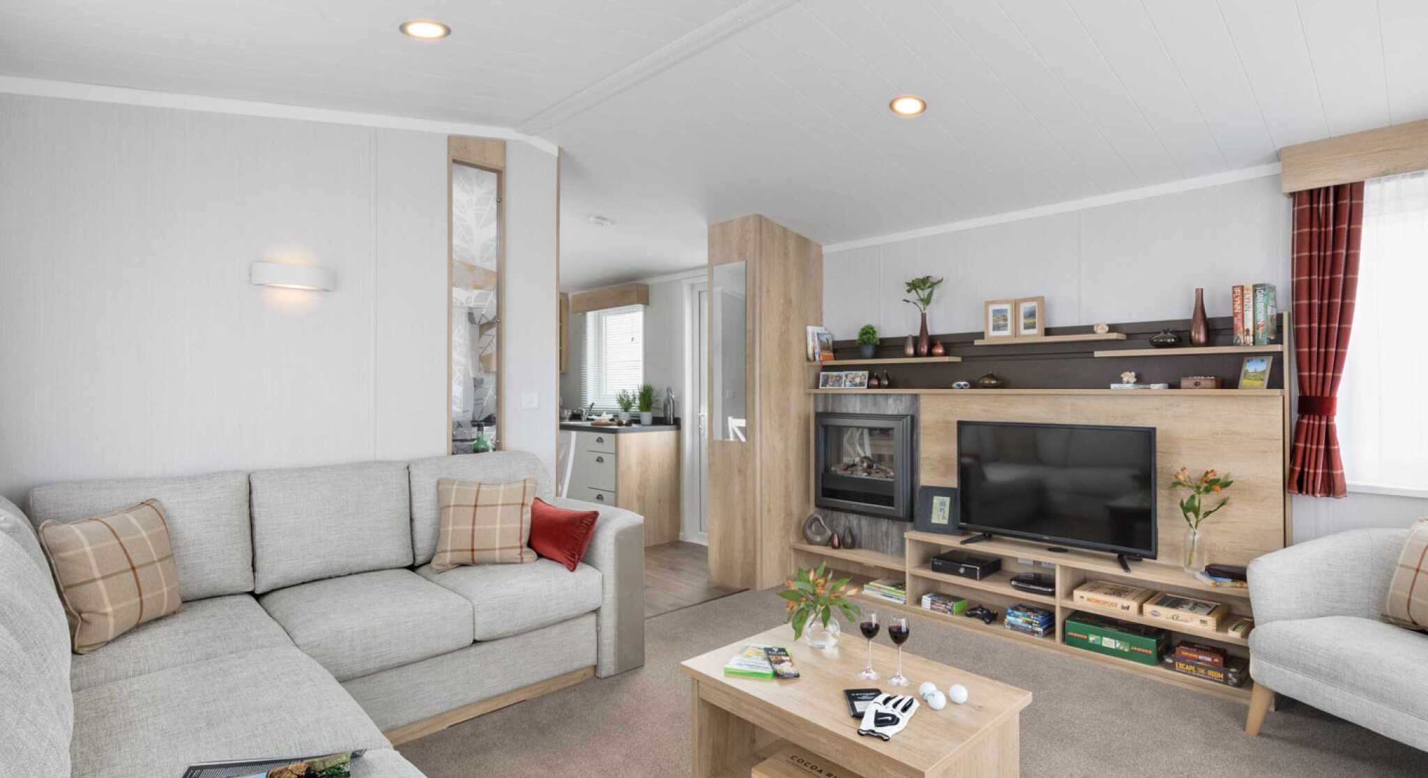 Inviting contemporary lounge featuring a grey corner sofa with patterned cushions, coffee table, TV, shelves, and soft, welcoming accents.
