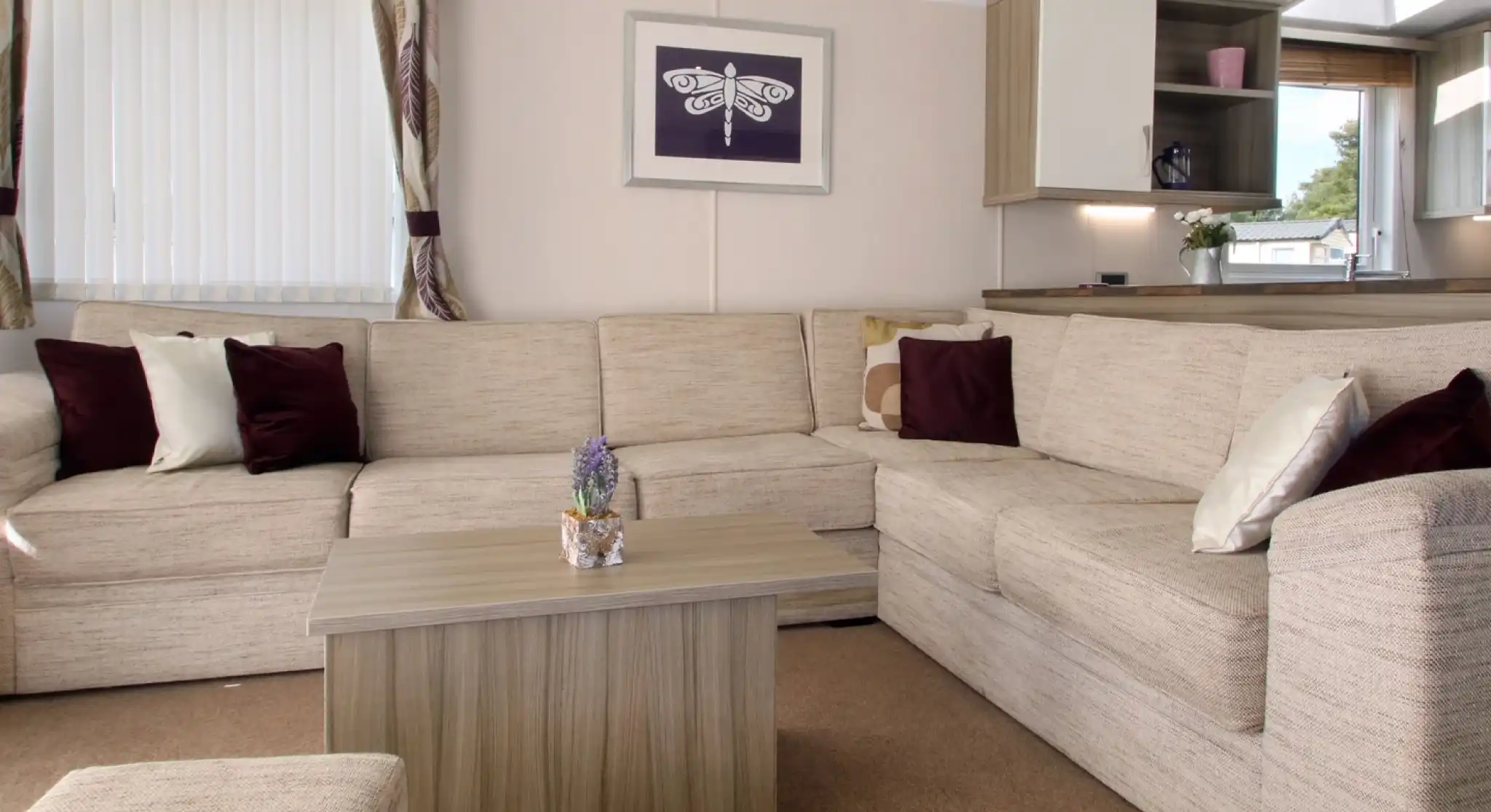 Cozy living room featuring a large beige sectional sofa, a small wooden coffee table with a decorative plant, and soft cushions in burgundy and cream. Natural light streams in through a window dressed with sheer curtains, complemented by a framed artwork on the wall.
