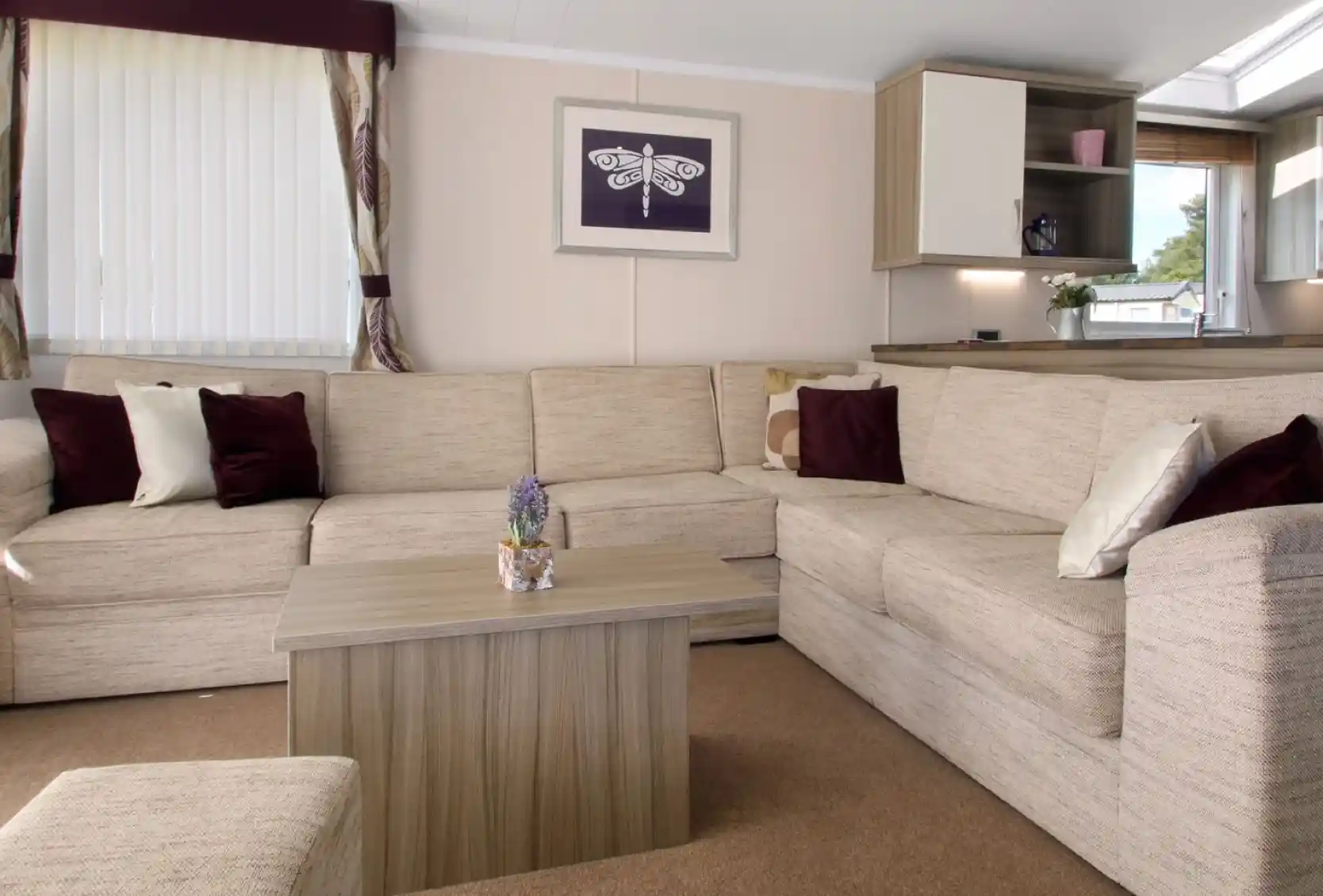 Cozy living room featuring a large beige sectional sofa, a small wooden coffee table with a decorative plant, and soft cushions in burgundy and cream. Natural light streams in through a window dressed with sheer curtains, complemented by a framed artwork on the wall.