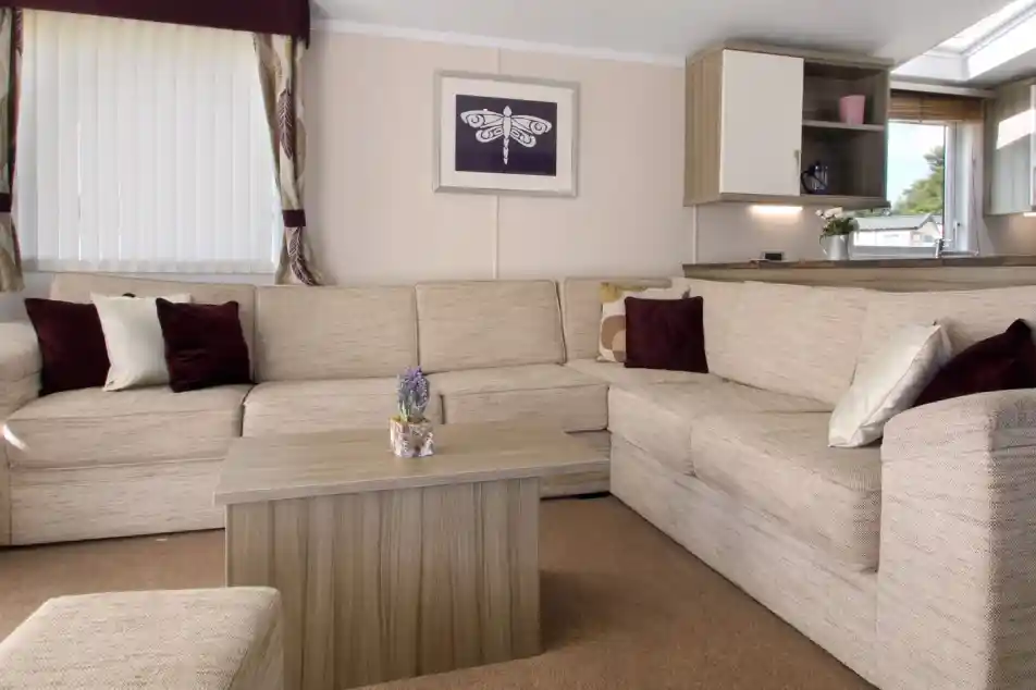 Cozy living room featuring a large beige sectional sofa, a small wooden coffee table with a decorative plant, and soft cushions in burgundy and cream. Natural light streams in through a window dressed with sheer curtains, complemented by a framed artwork on the wall.