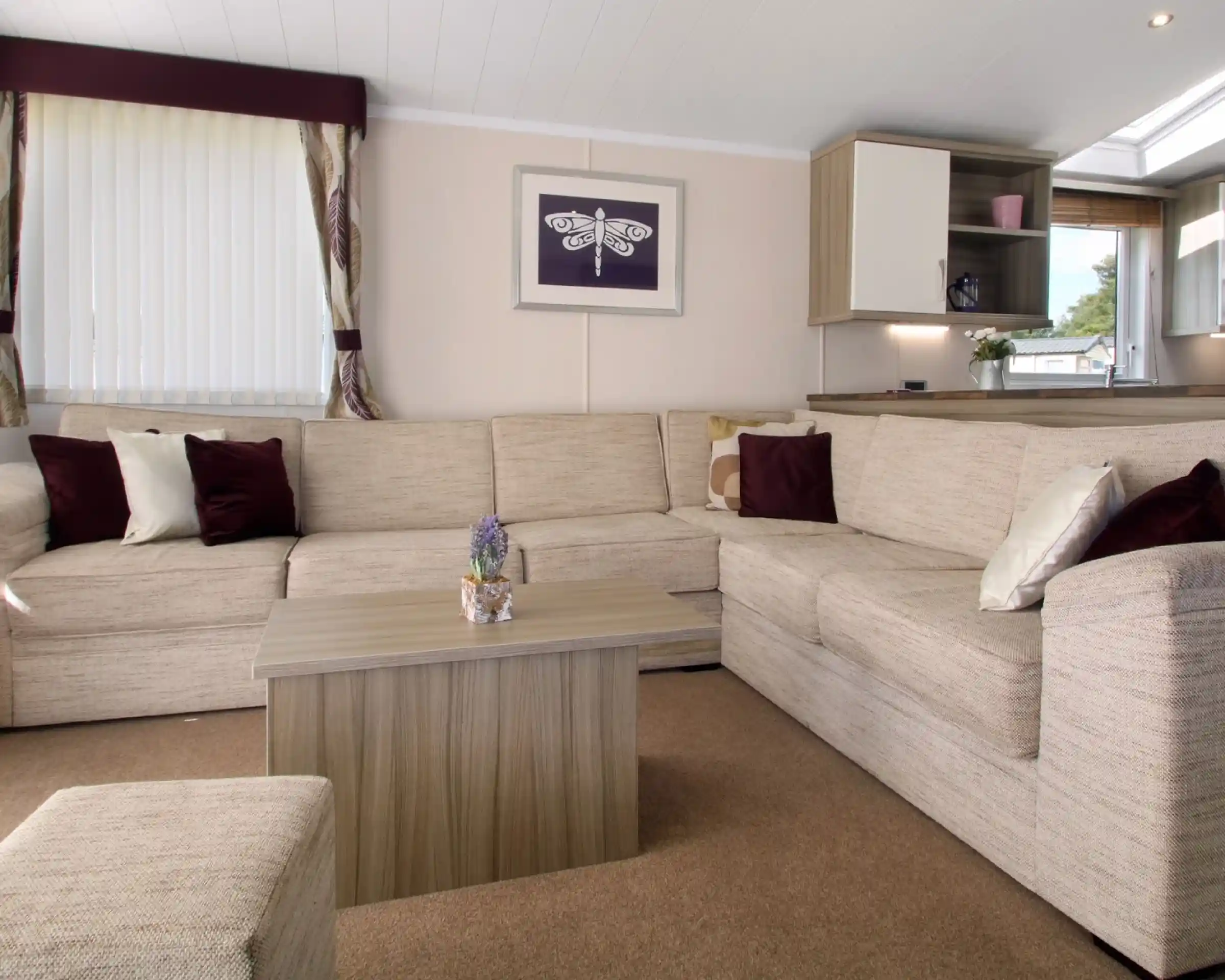 Cozy living room featuring a large beige sectional sofa, a small wooden coffee table with a decorative plant, and soft cushions in burgundy and cream. Natural light streams in through a window dressed with sheer curtains, complemented by a framed artwork on the wall.