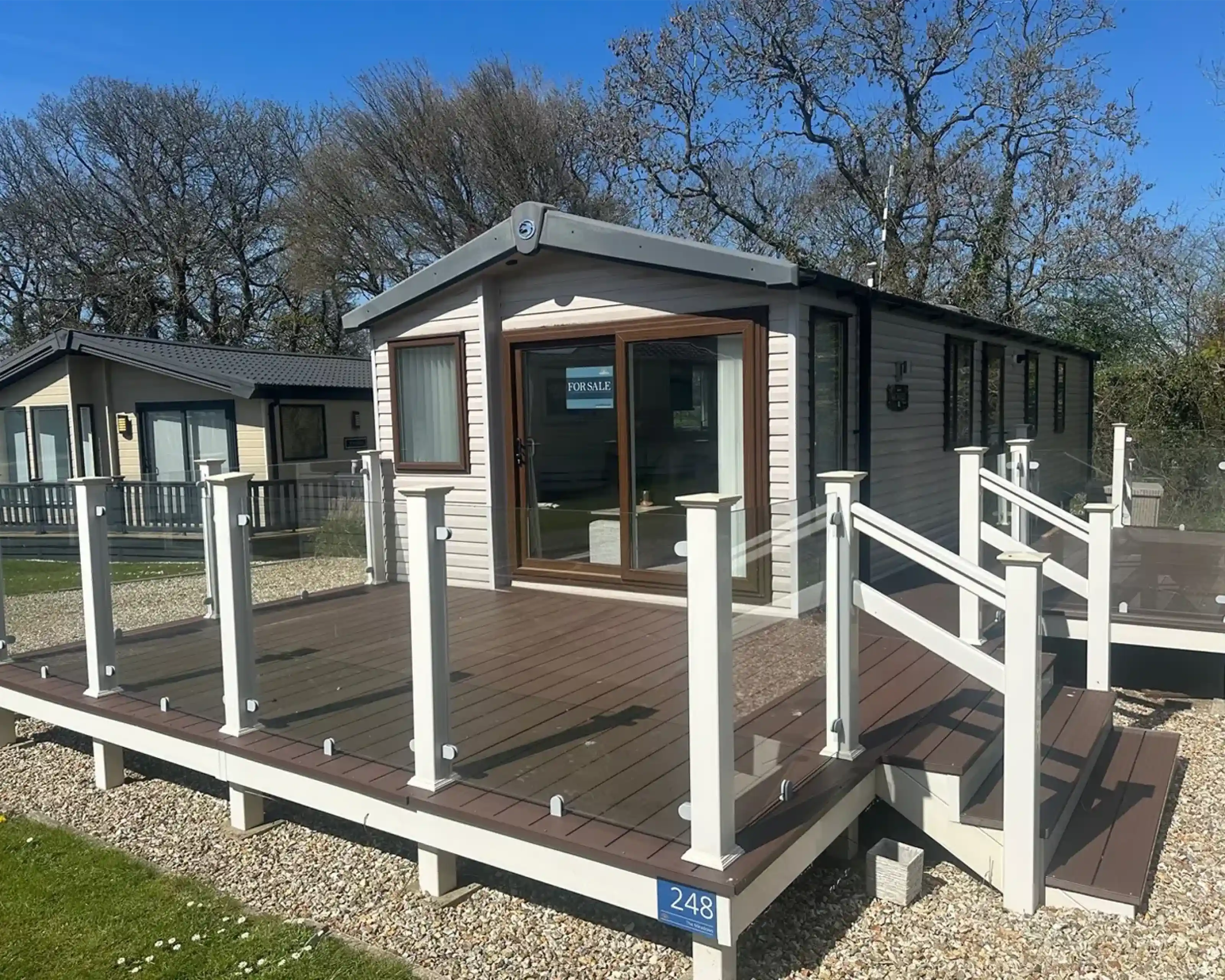A modern mobile home with large windows and a deck featuring glass railings. The surrounding area includes grassy patches and trees, creating a serene environment. A "For Sale" sign is visible in one window.