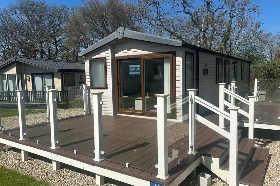 A modern mobile home with large windows and a deck featuring glass railings. The surrounding area includes grassy patches and trees, creating a serene environment. A "For Sale" sign is visible in one window.