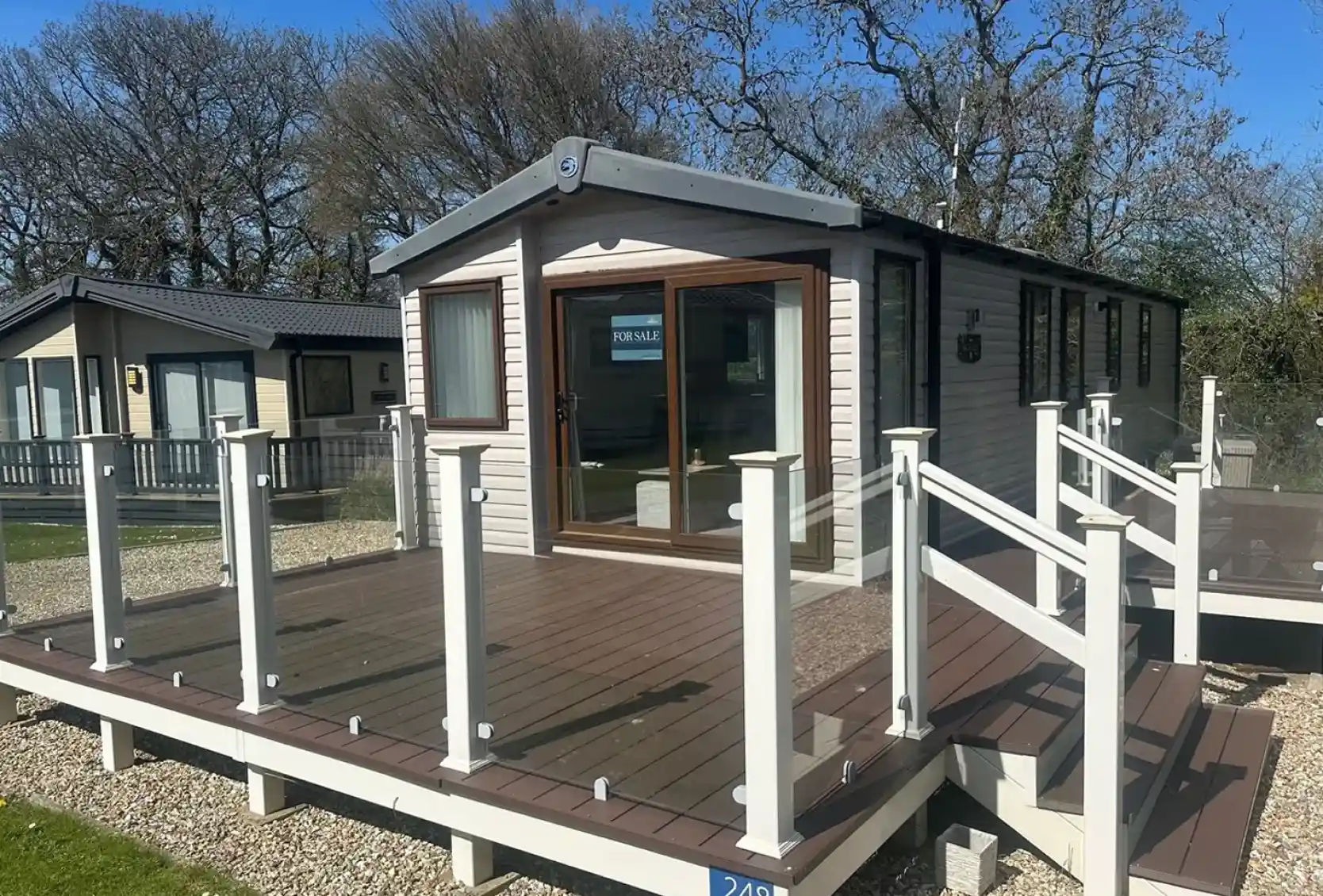 A modern mobile home with large windows and a deck featuring glass railings. The surrounding area includes grassy patches and trees, creating a serene environment. A "For Sale" sign is visible in one window.