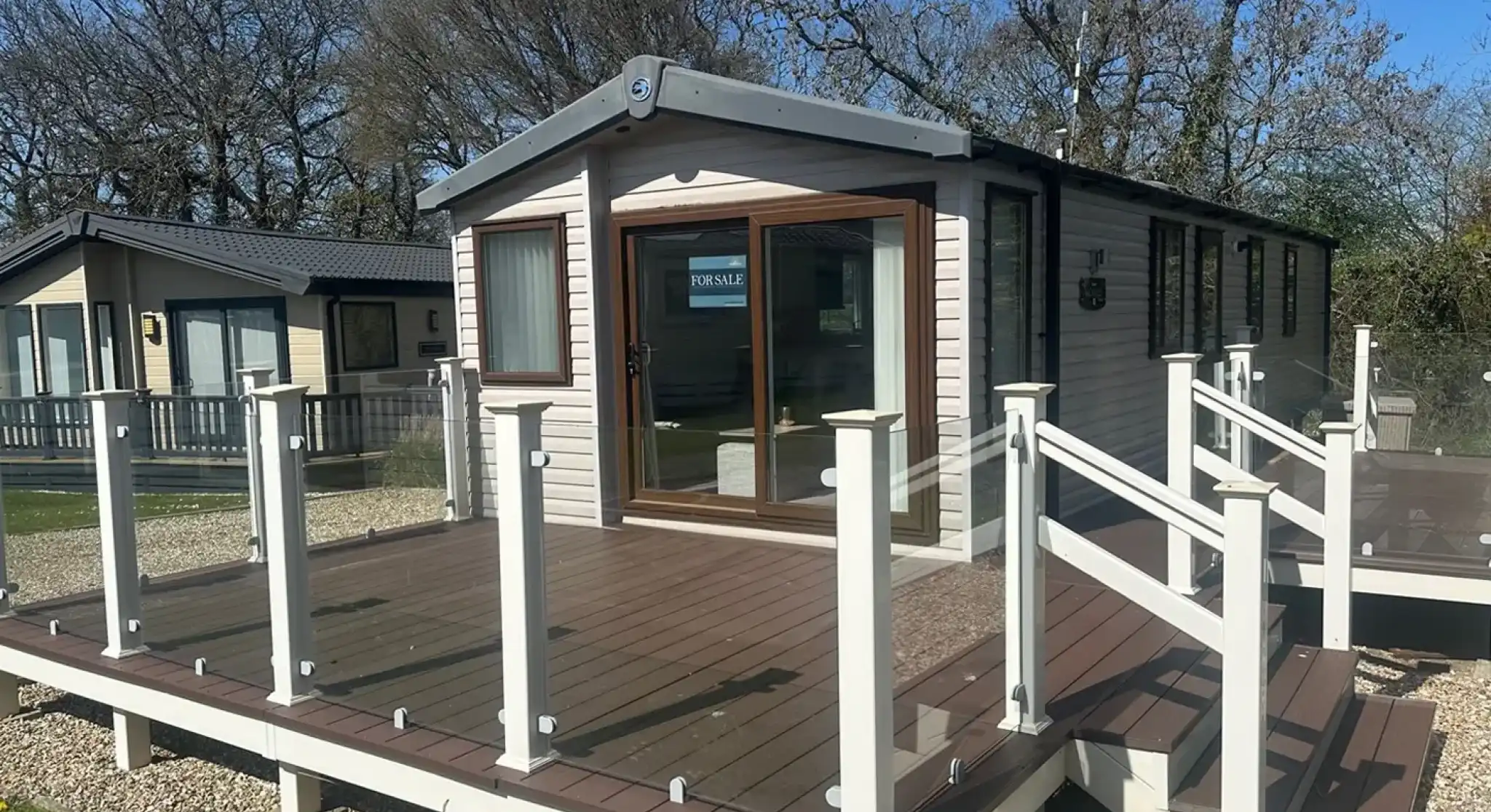 A modern mobile home with large windows and a deck featuring glass railings. The surrounding area includes grassy patches and trees, creating a serene environment. A "For Sale" sign is visible in one window.
