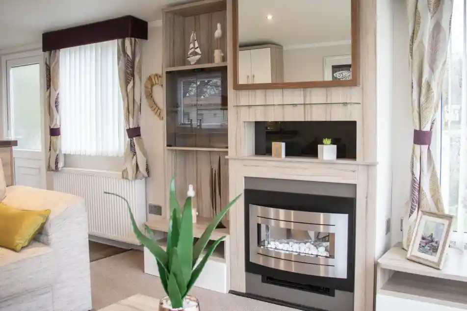A cozy living room with a light color scheme features a modern fireplace with a mirror above it. There are built-in shelves with decorative items and a potted plant on the coffee table. Natural light streams in through the window, which has white blinds. A comfortable sofa adds to the inviting atmosphere.