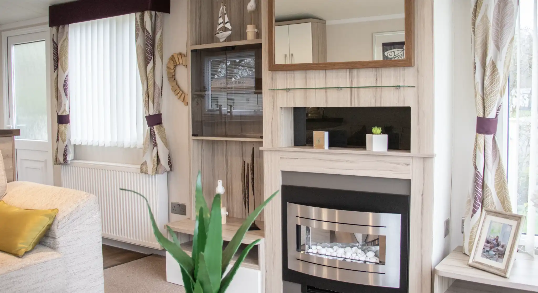 A cozy living room with a light color scheme features a modern fireplace with a mirror above it. There are built-in shelves with decorative items and a potted plant on the coffee table. Natural light streams in through the window, which has white blinds. A comfortable sofa adds to the inviting atmosphere.